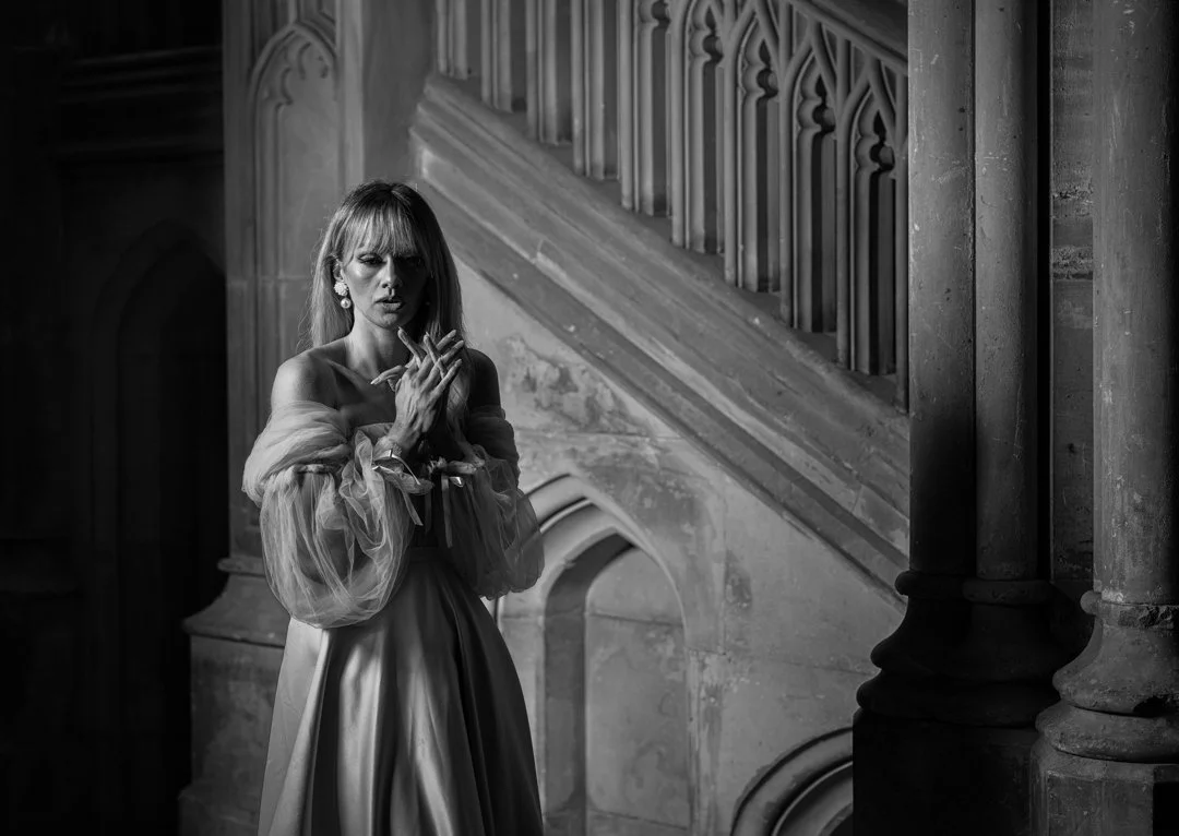 A woman in a vintage gown standing on a staircase inside a gothic-style building, black and white photo.
