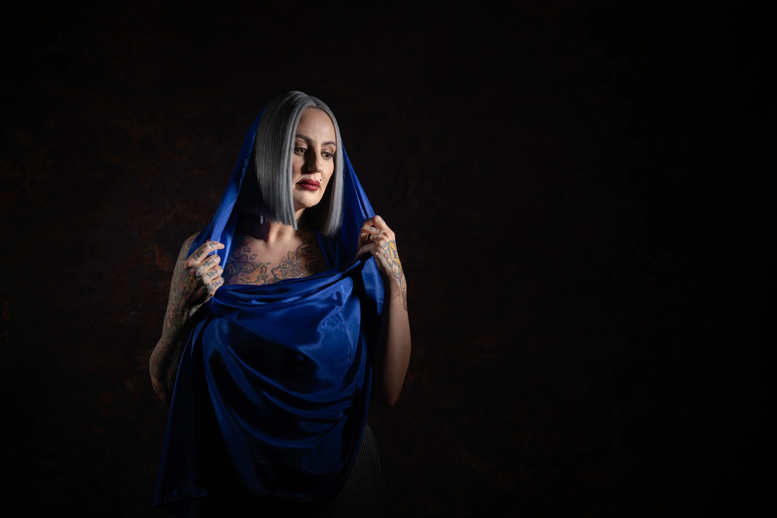 Woman with a gray bob haircut and tattoos on her arms and chest, holding a blue satin fabric against her body, against a dark background.
