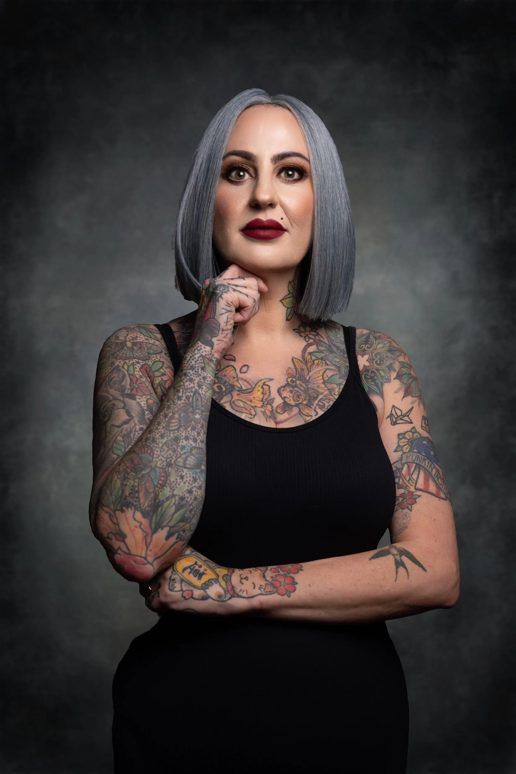 A woman with shoulder-length gray hair, tattoos on her arms and chest, wearing a black sleeveless top, standing with her hand near her chin against a dark, textured background.