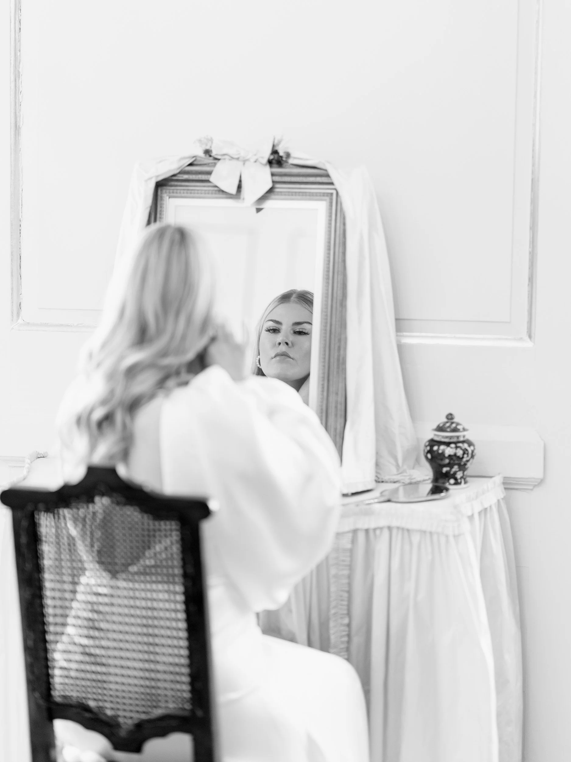 A woman with wavy hair looking at her reflection in a mirror on a dresser, with a decorative container beside it, in black and white.