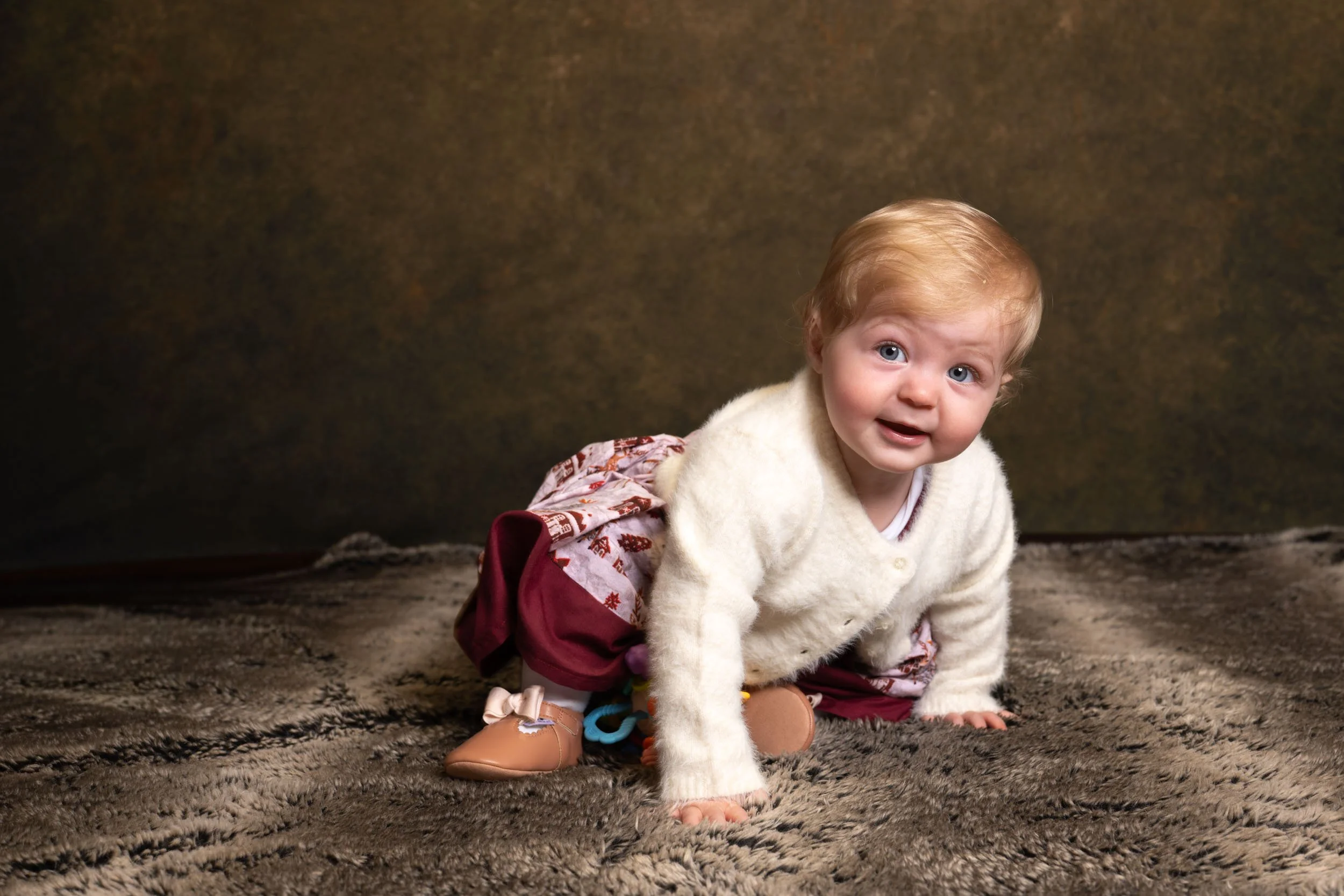 A young child crawling on a brown shaggy carpet, wearing a cream fuzzy sweater, maroon pants with a pattern, pink shoes, and has light brown hair and blue eyes.