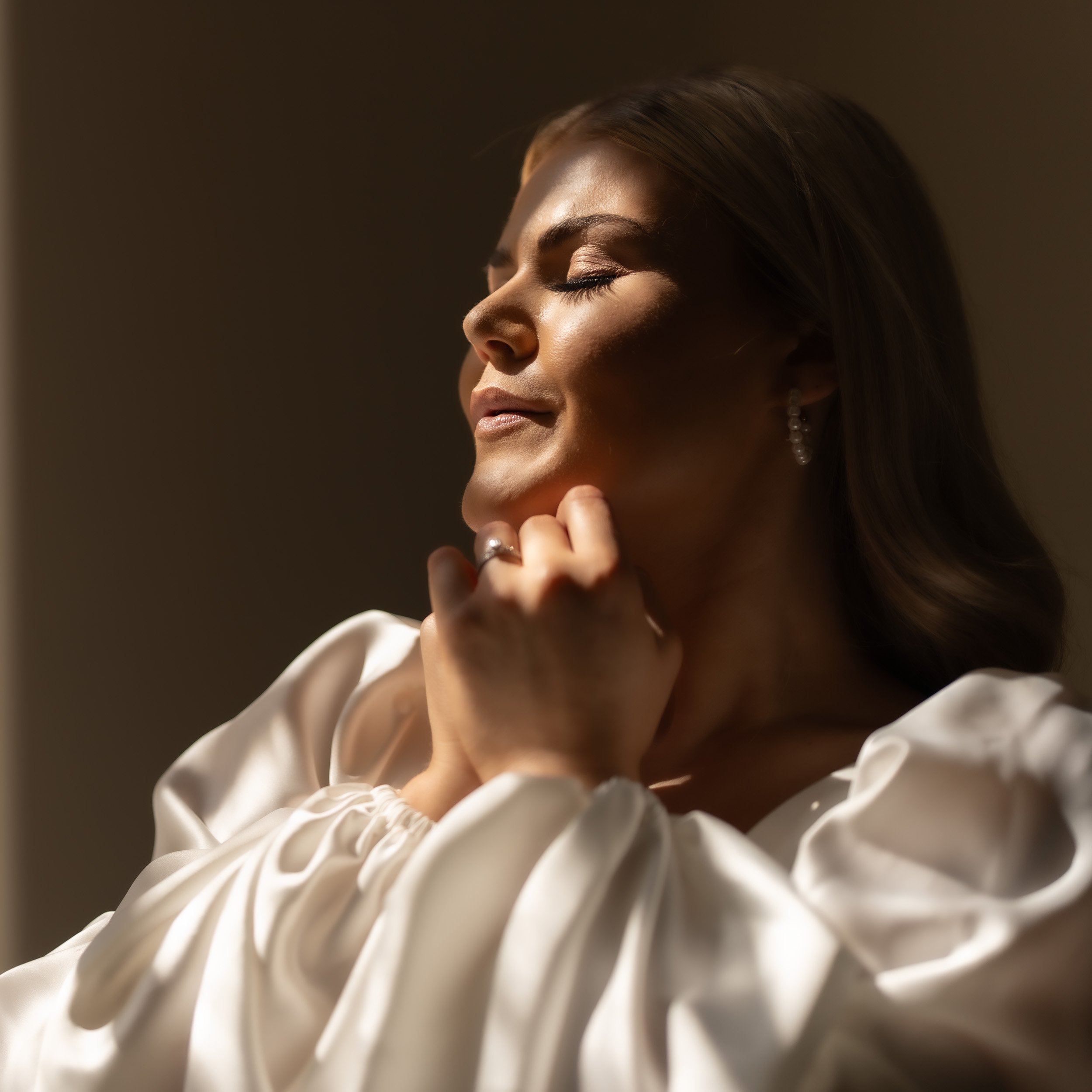 A woman with closed eyes and a peaceful expression, wearing earrings and a ring, in soft lighting.