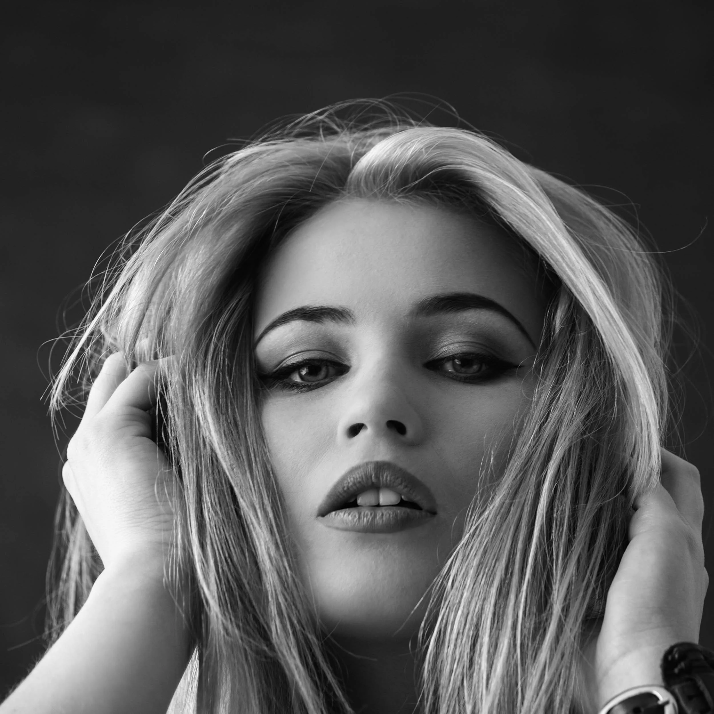 Black and white close-up portrait of a woman with long, tousled hair, holding her hair with both hands, looking directly at the camera.