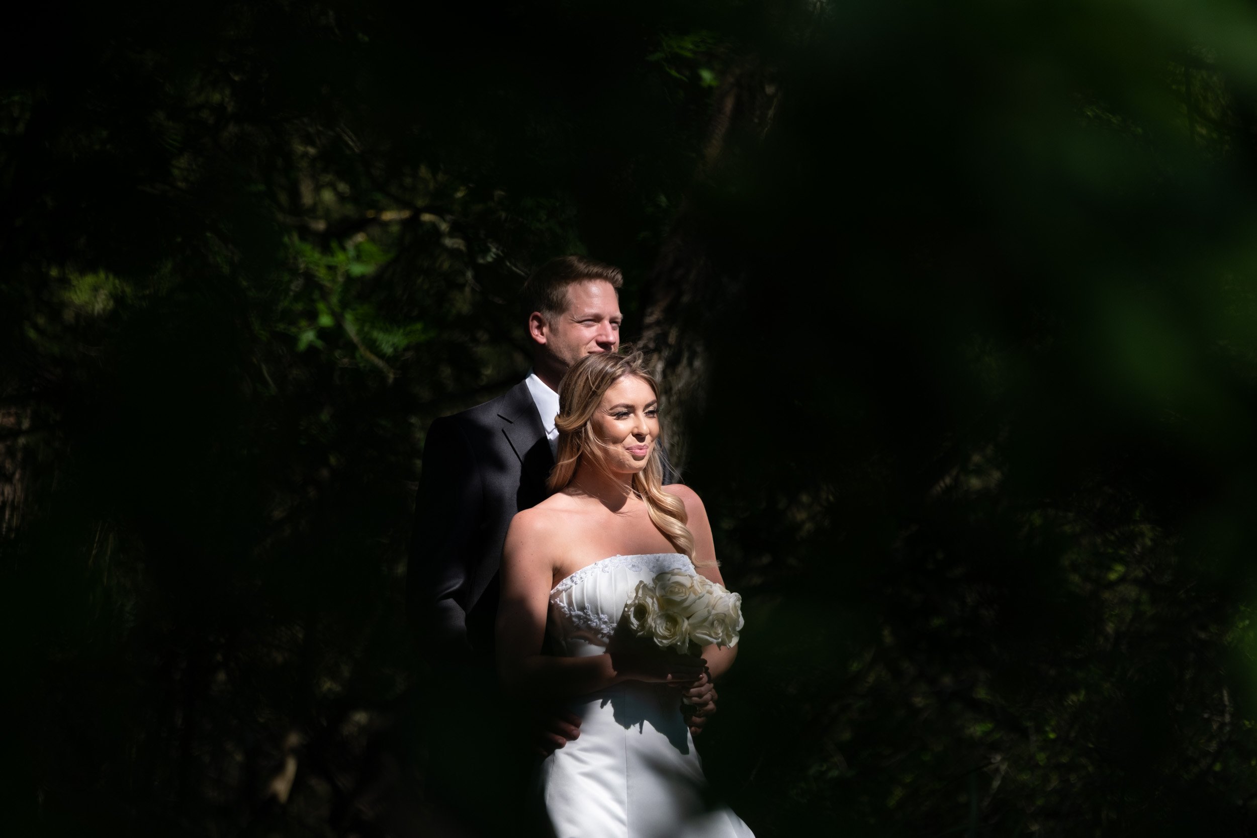 A bride and groom standing together outdoors in the woods, with the bride holding a bouquet of white roses, in a moment of quiet happiness.
