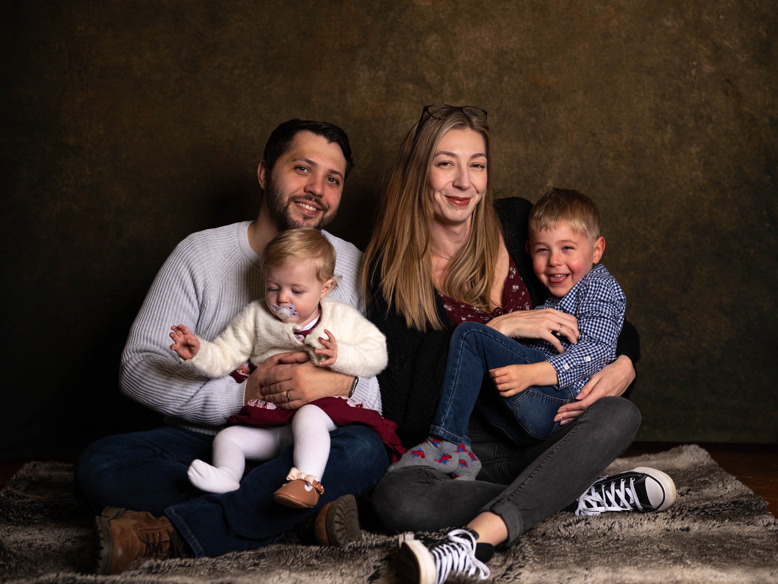 A family of four sitting on a blanket, smiling, in a studio with a dark background. The father has short dark hair and a beard, holding a little girl with a pacifier and white tights. The mother has long blonde hair and is sitting next to the father,