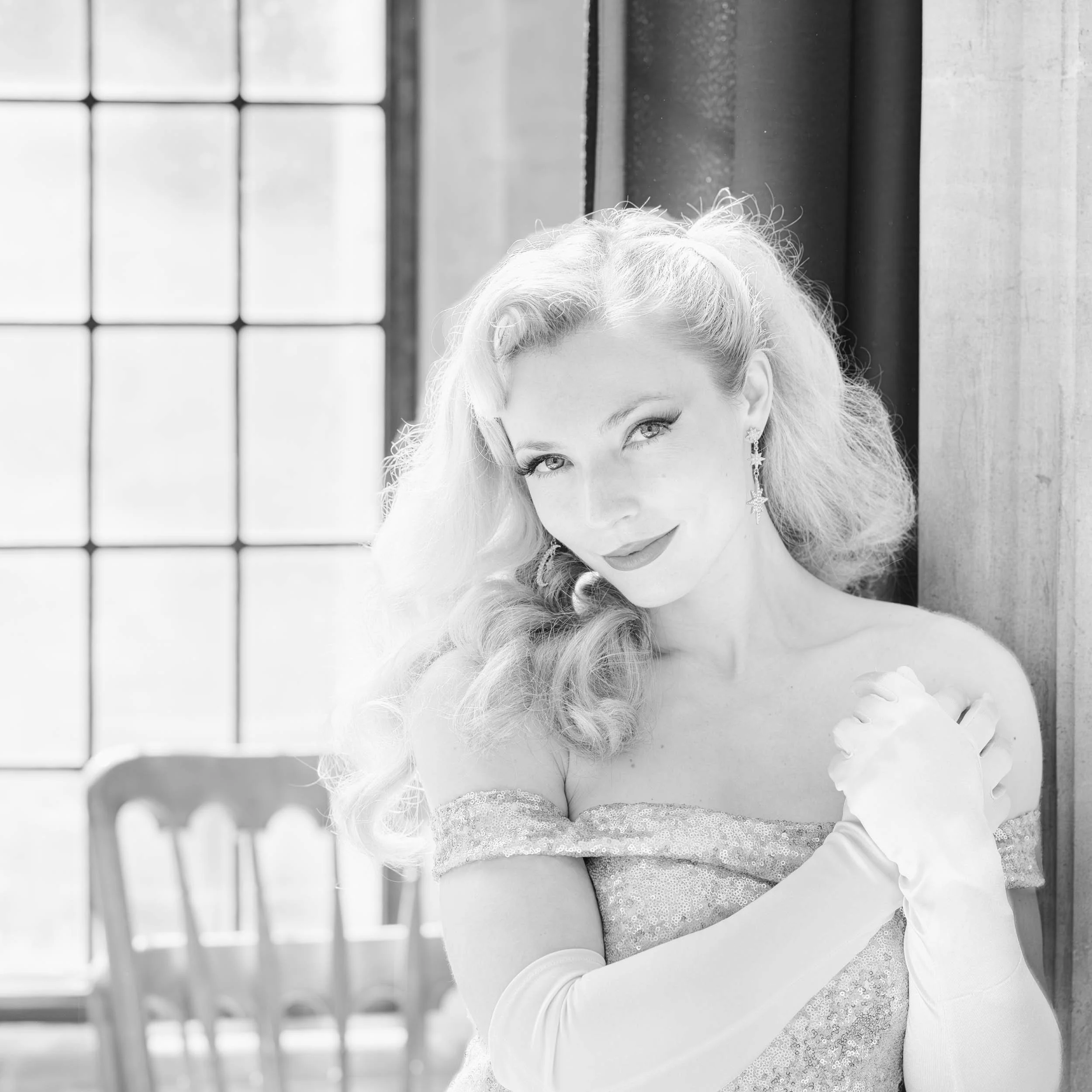 Black and white photo of a woman with vintage style hair and makeup, wearing long gloves and earrings, standing near a window with sunlight streaming in.
