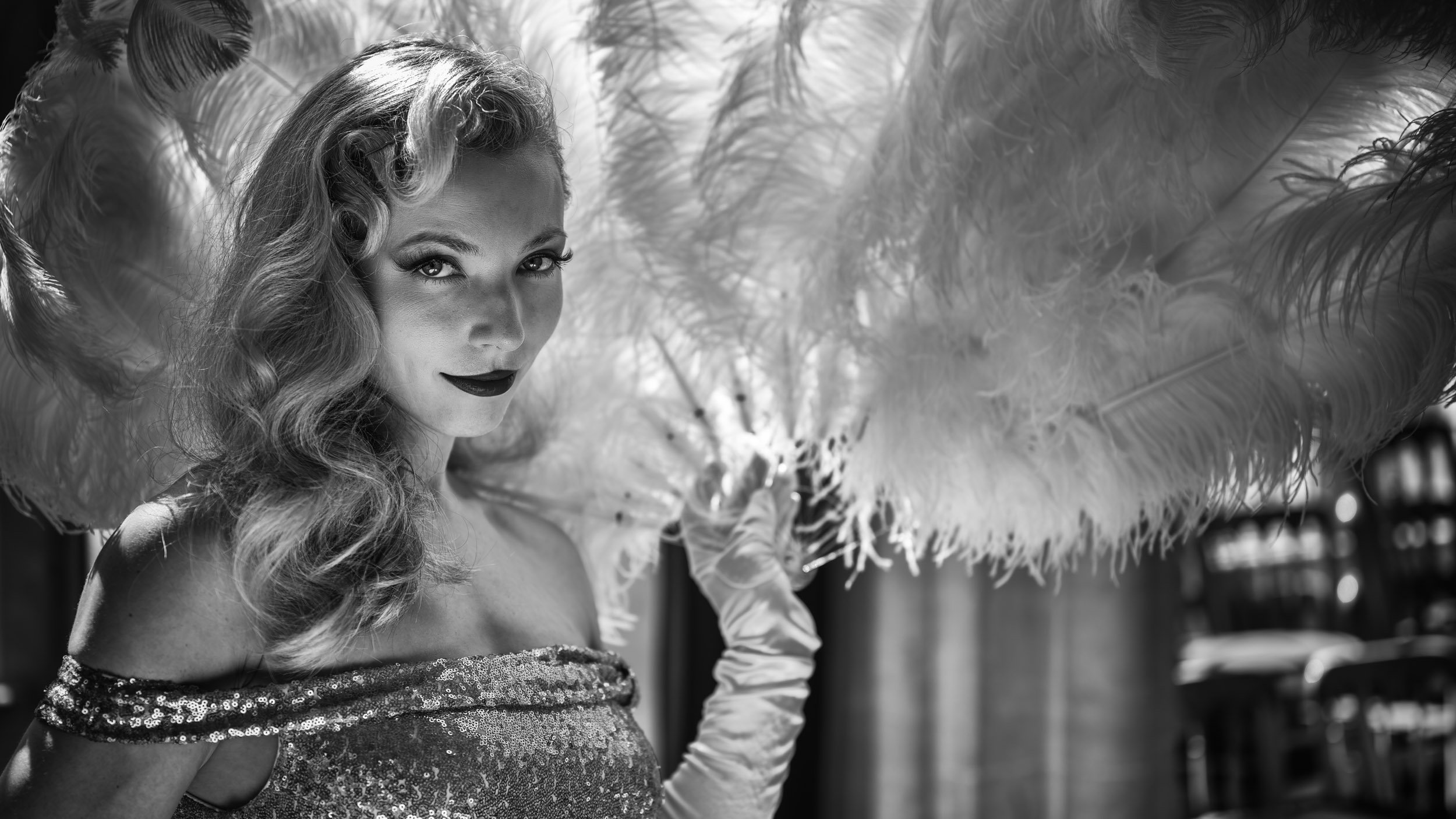 A woman with wavy hair, wearing a sequined off-shoulder dress, holding a feathered fan in an outdoor setting.