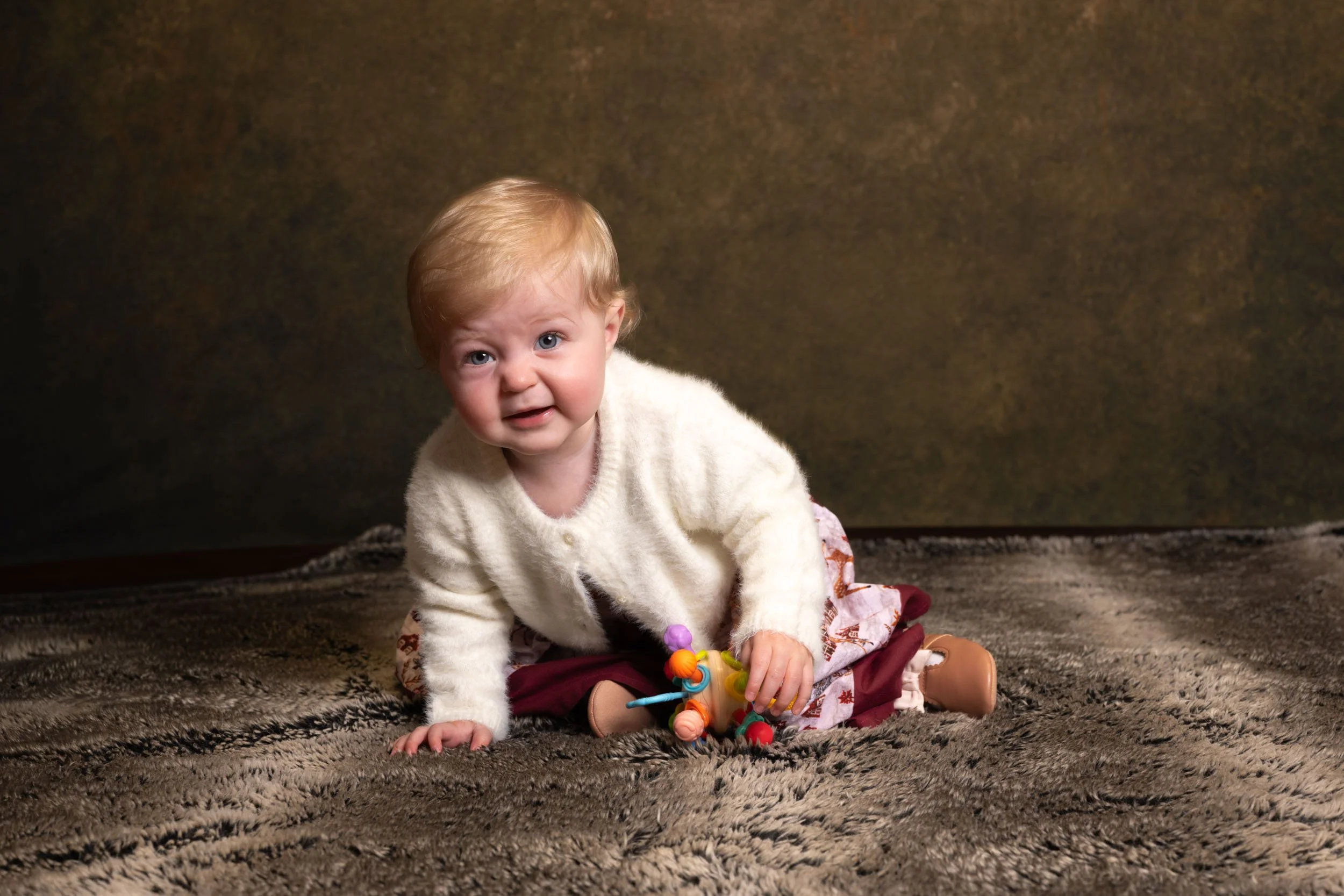 A young child with blonde hair and blue eyes sitting on a patterned brown carpet, wearing a white sweater and holding a colorful toy, looking at the camera.
