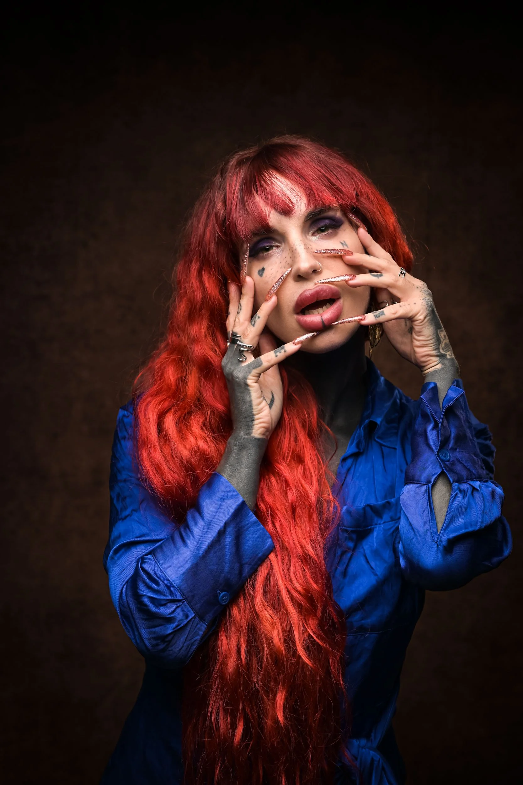 Woman with long red curly hair and tattoos on her hands and face, wearing a blue shirt, posing against a dark background.