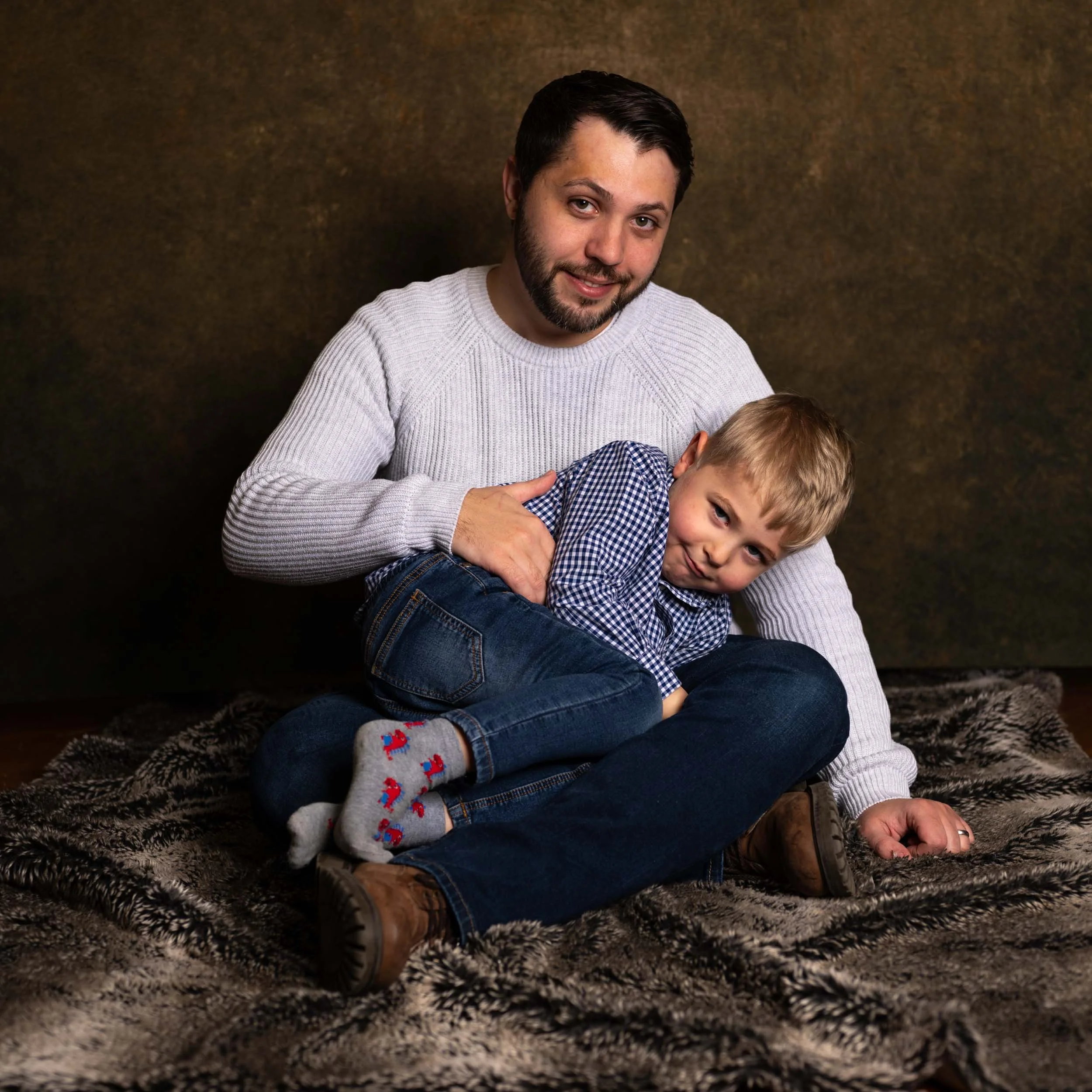 A man and young boy sitting on a patterned blanket, with the man smiling and the boy making a playful face, in a studio setting.