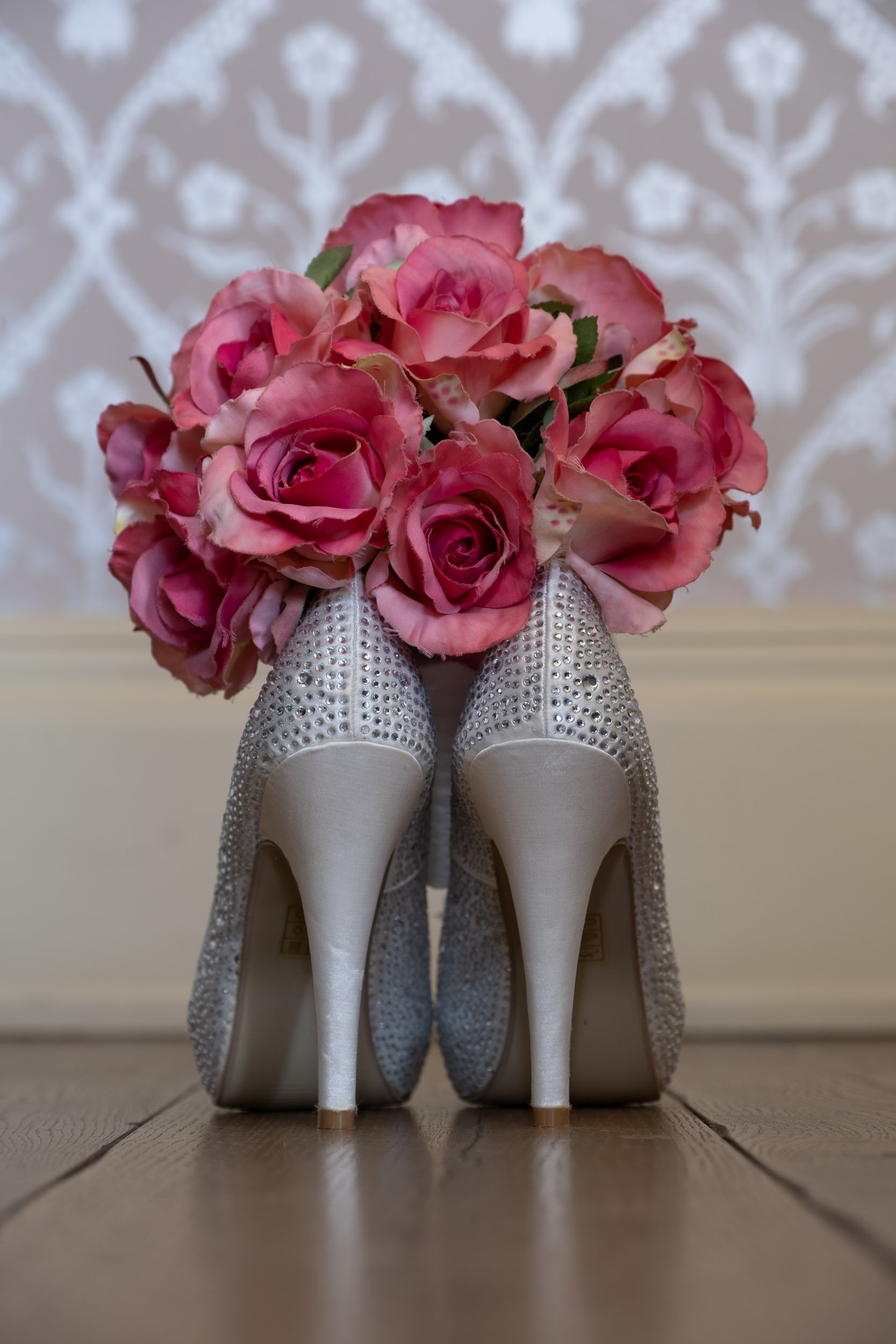 A bouquet of pink roses and lilies placed on top of a pair of high-heeled shoes decorated with silver rhinestones, with a background of damask patterned wallpaper.