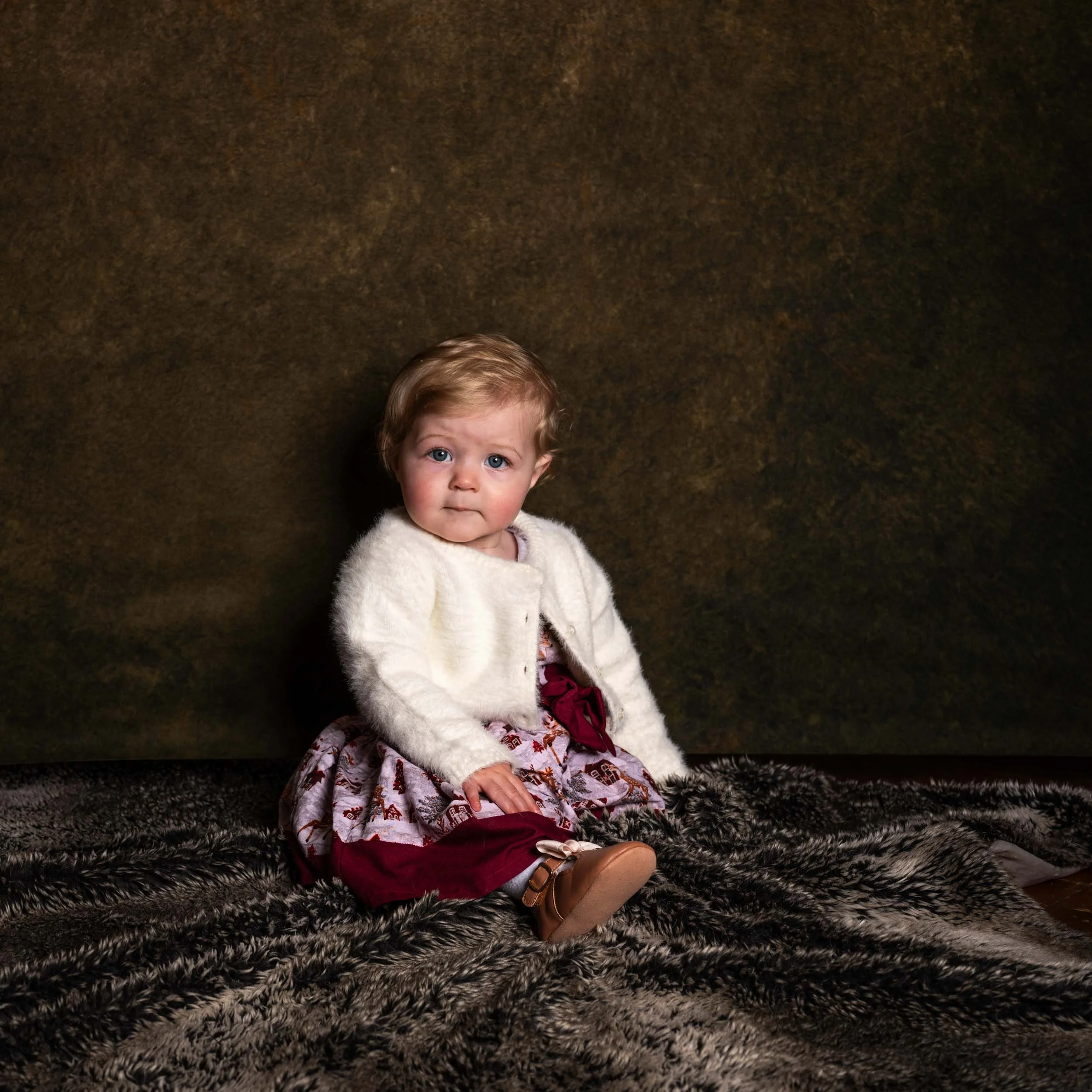 A young girl with curly blonde hair, blue eyes, and a serious expression, sitting on a textured fur blanket against a brown background, wearing a white fuzzy cardigan, patterned dress, and brown shoes.