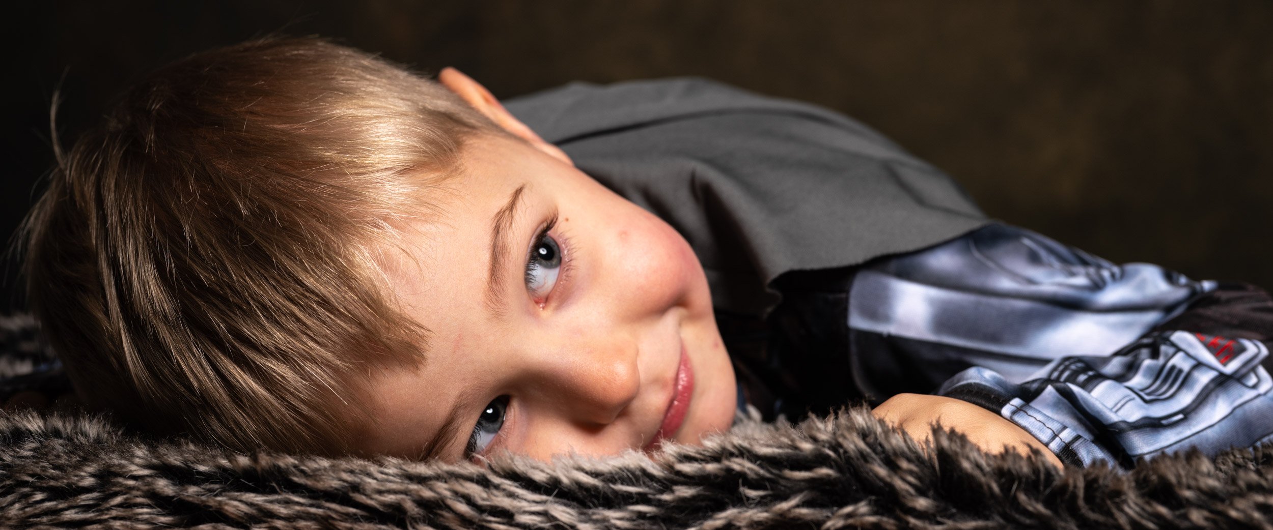 Close-up of a young boy with blonde hair and blue eyes lying on a furry surface, looking up with a slight smile.