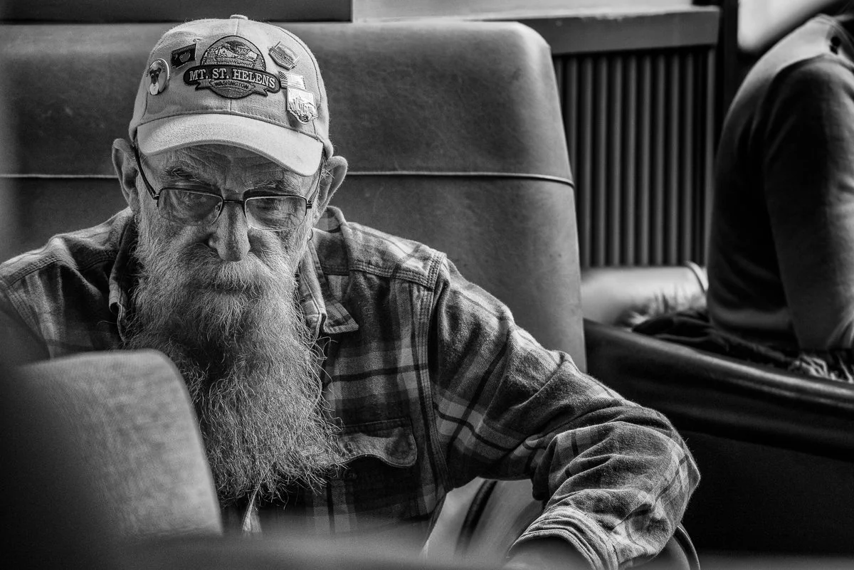 An elderly man with a long beard and glasses, wearing a cap with patches, sitting in a restaurant or cafe.