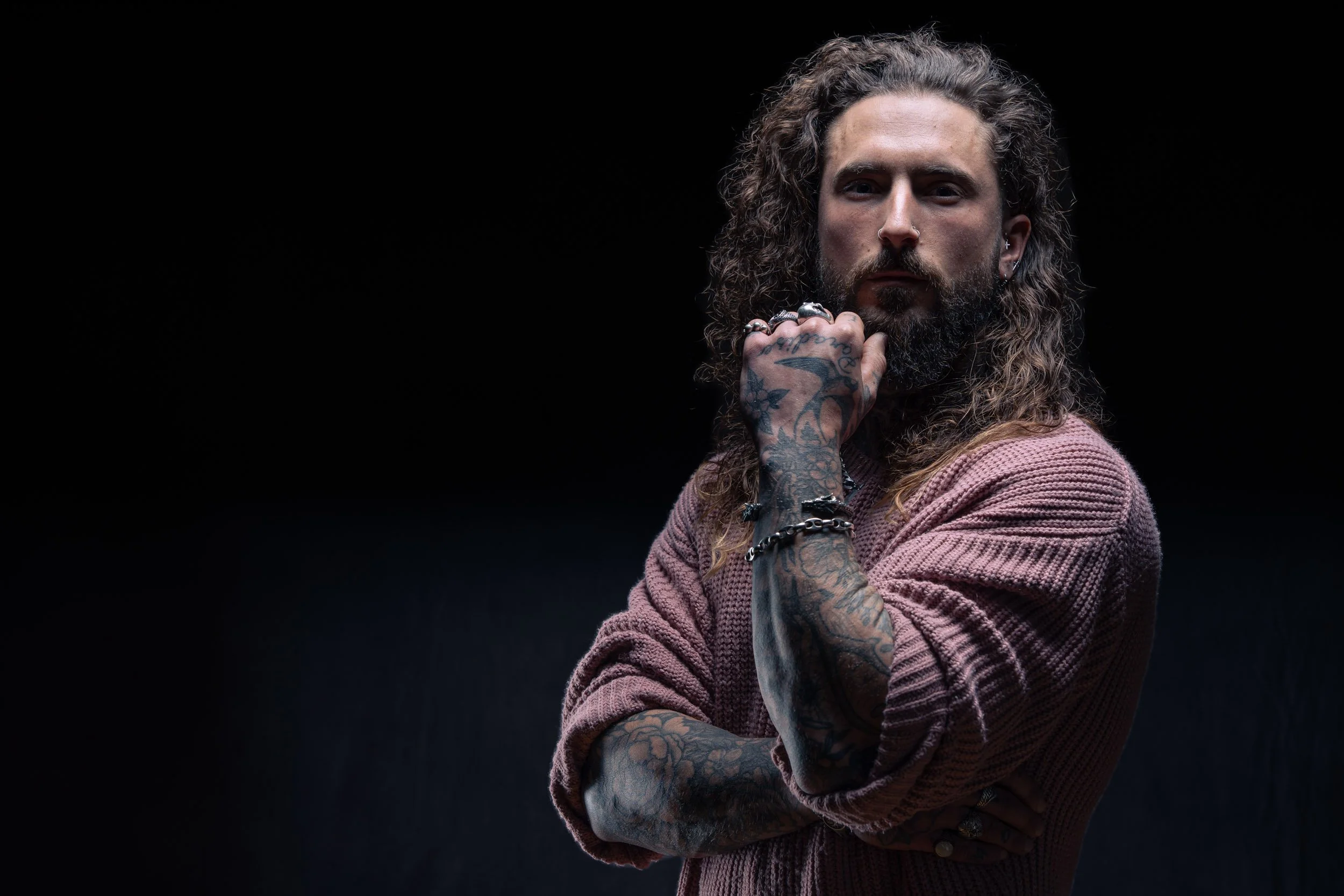 Portrait of a man with long curly hair and tattoos on his arms, wearing a pink sweater, posing with his chin resting on his hand against a dark background.