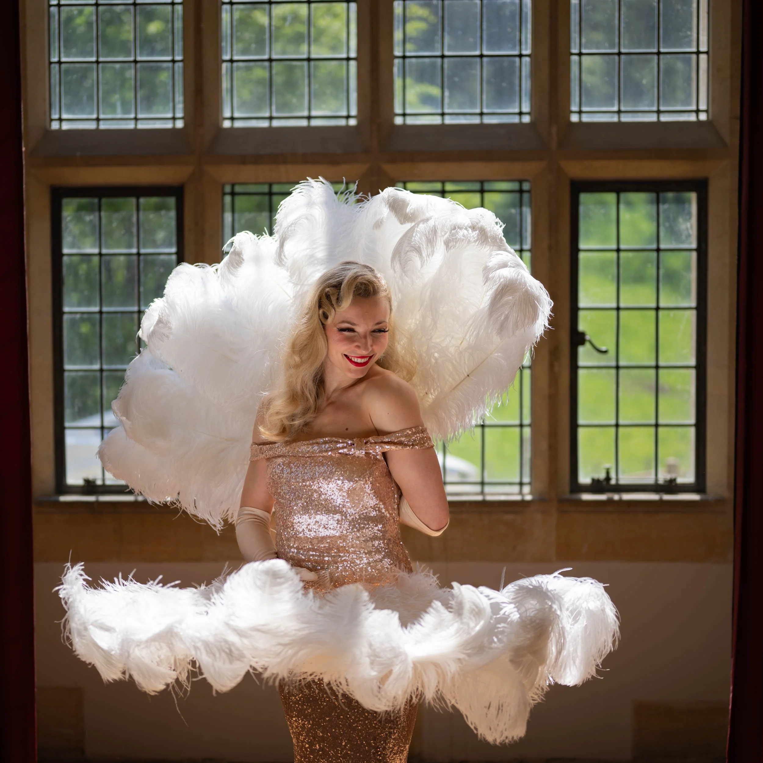 A woman in a sparkling gold dress with large white feathered wings standing in front of large windows, smiling and winking.