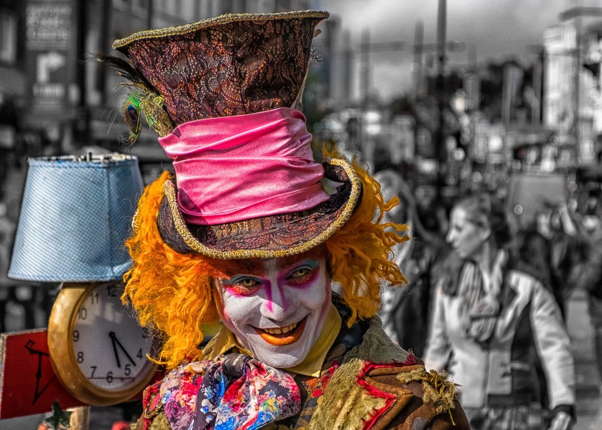 A person dressed as a clown with bright orange curly hair, a painted white face with pink and purple makeup, wearing a colorful patchwork costume and a tall, elaborate hat with pink satin and patterned fabric, standing in a busy street with blurred pedestrians in the background.