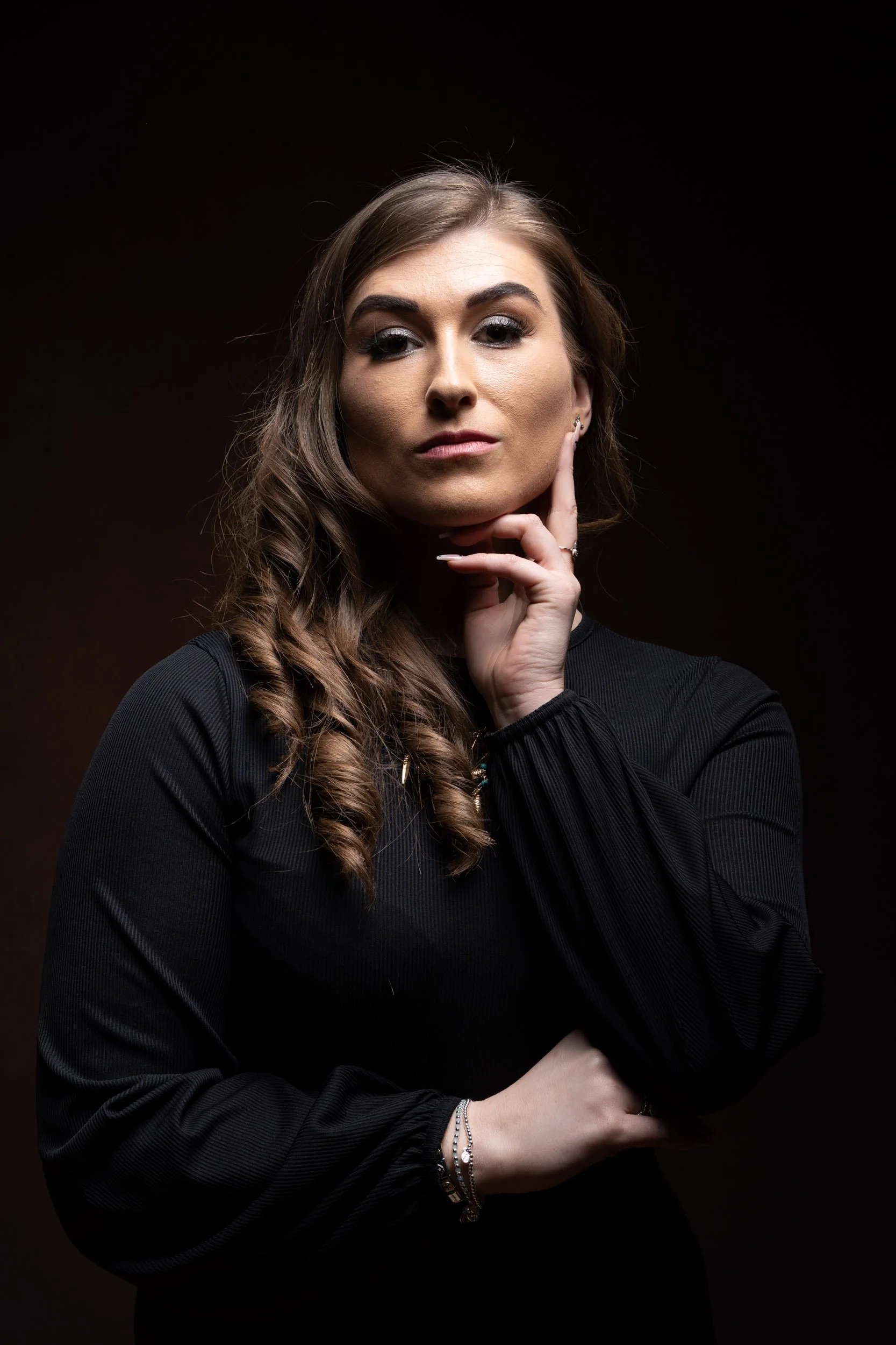 Portrait of a serious woman with long wavy brown hair, wearing a black long-sleeve top, posing thoughtfully with her hand near her chin against a dark background.