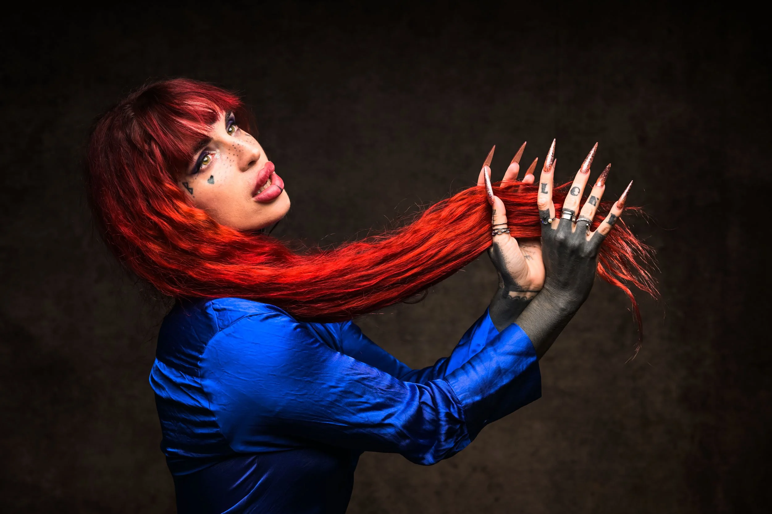 Woman with red hair and tattoos, wearing black gloves and a blue shirt, holding and examining long, pointed, metallic nails or claws.