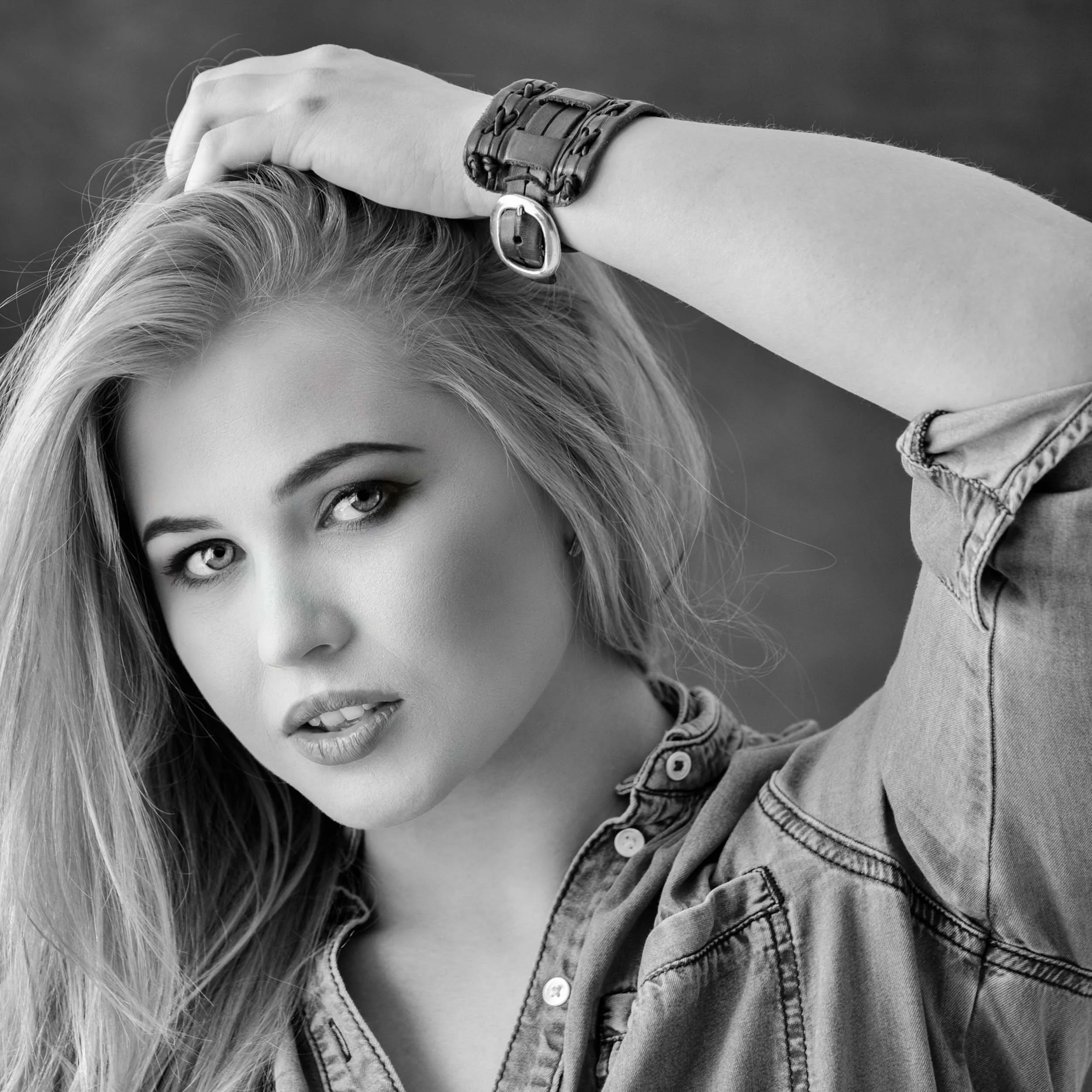 Black and white photo of a young woman with long blonde hair, looking directly at the camera, wearing a denim shirt and a wristwatch with her arm resting on her head.
