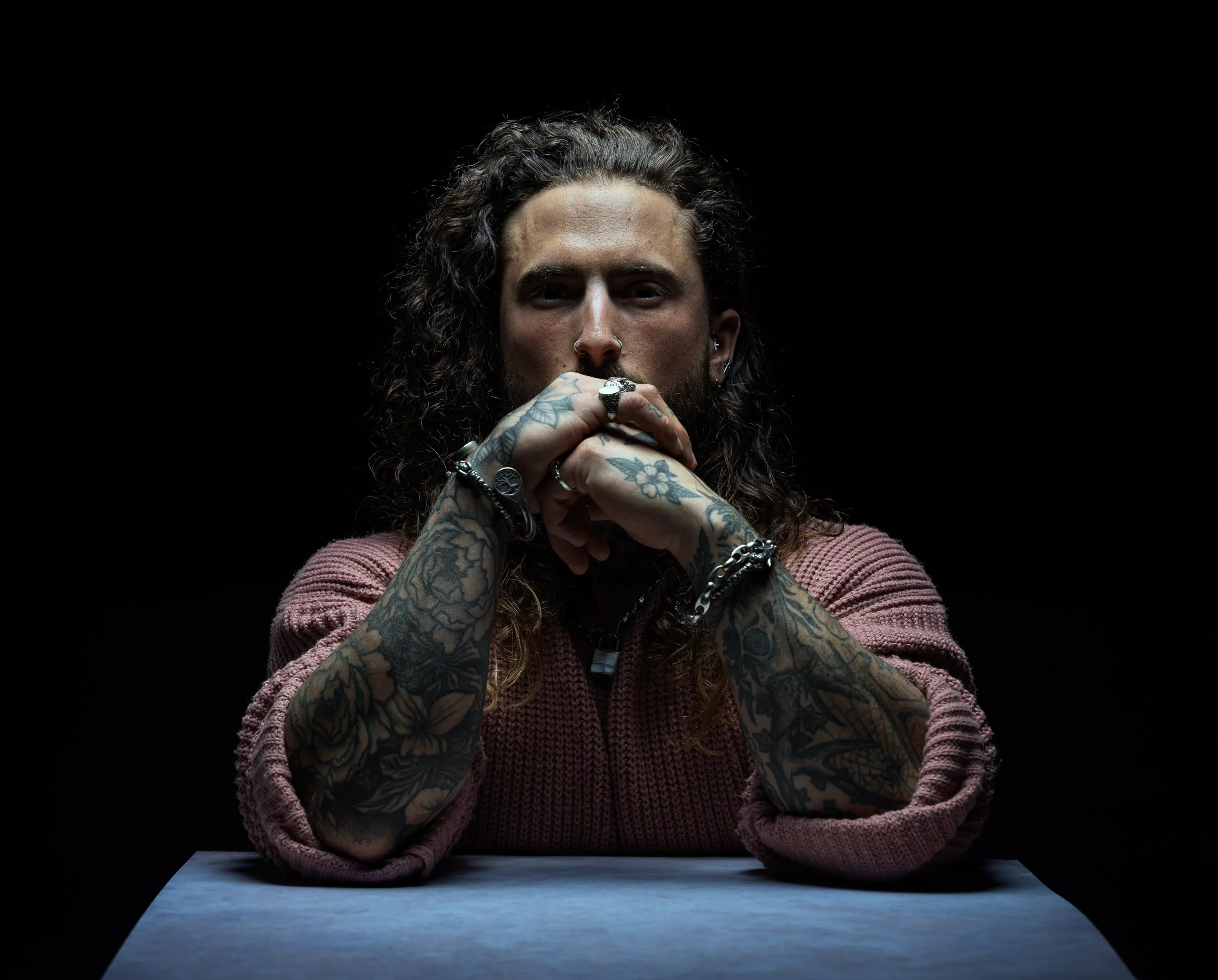 A man with long, curly hair and tattoos on both arms is sitting at a table with his hands clasped in front of his face, looking directly at the camera against a black background.