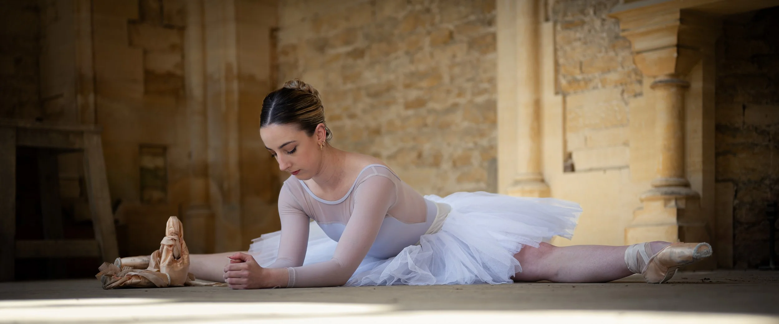 Ballet dancer in a white tutu stretching on a wooden floor in a rustic studio, with a brick wall in the background.