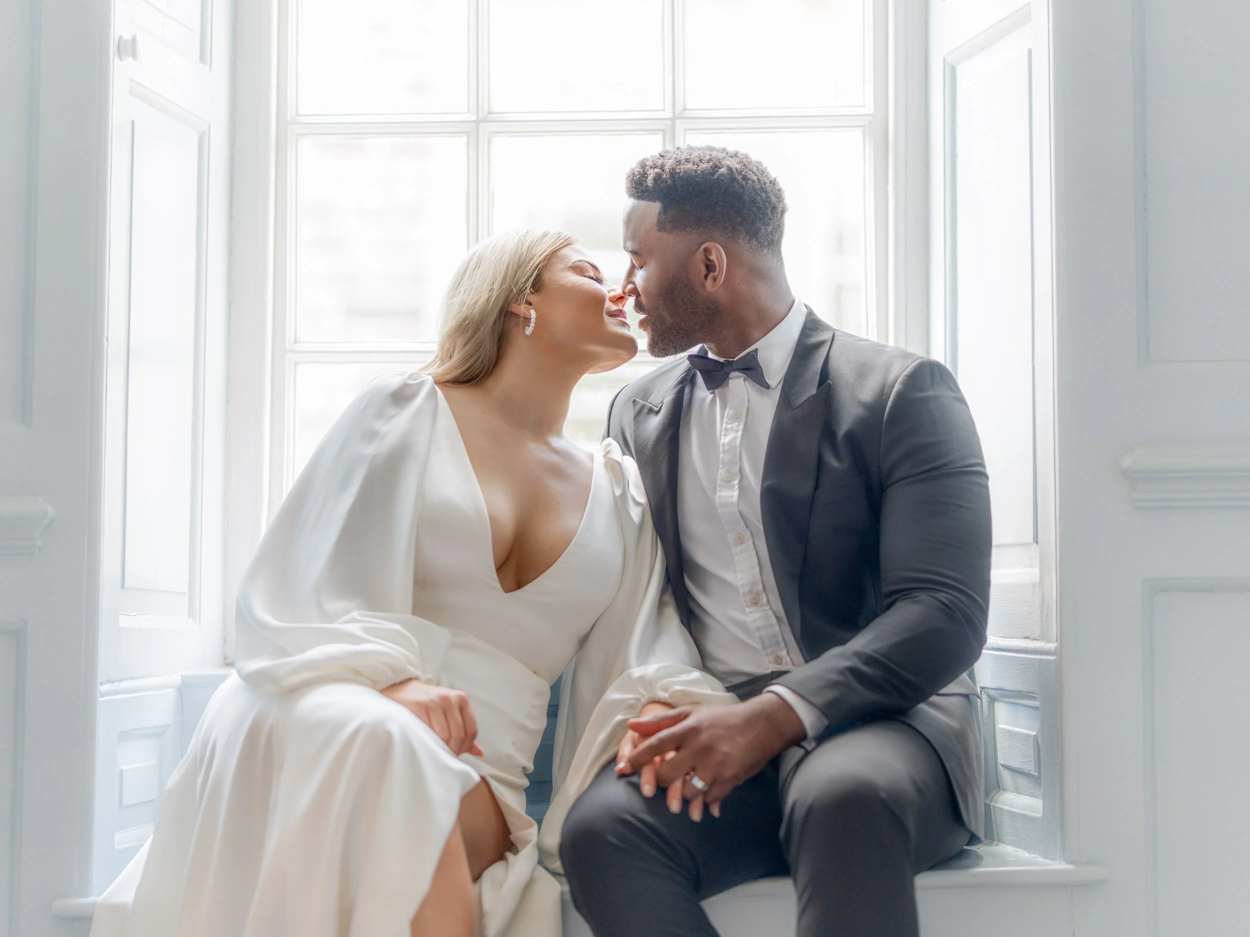 A couple dressed in wedding attire sharing an intimate moment by a bright window.