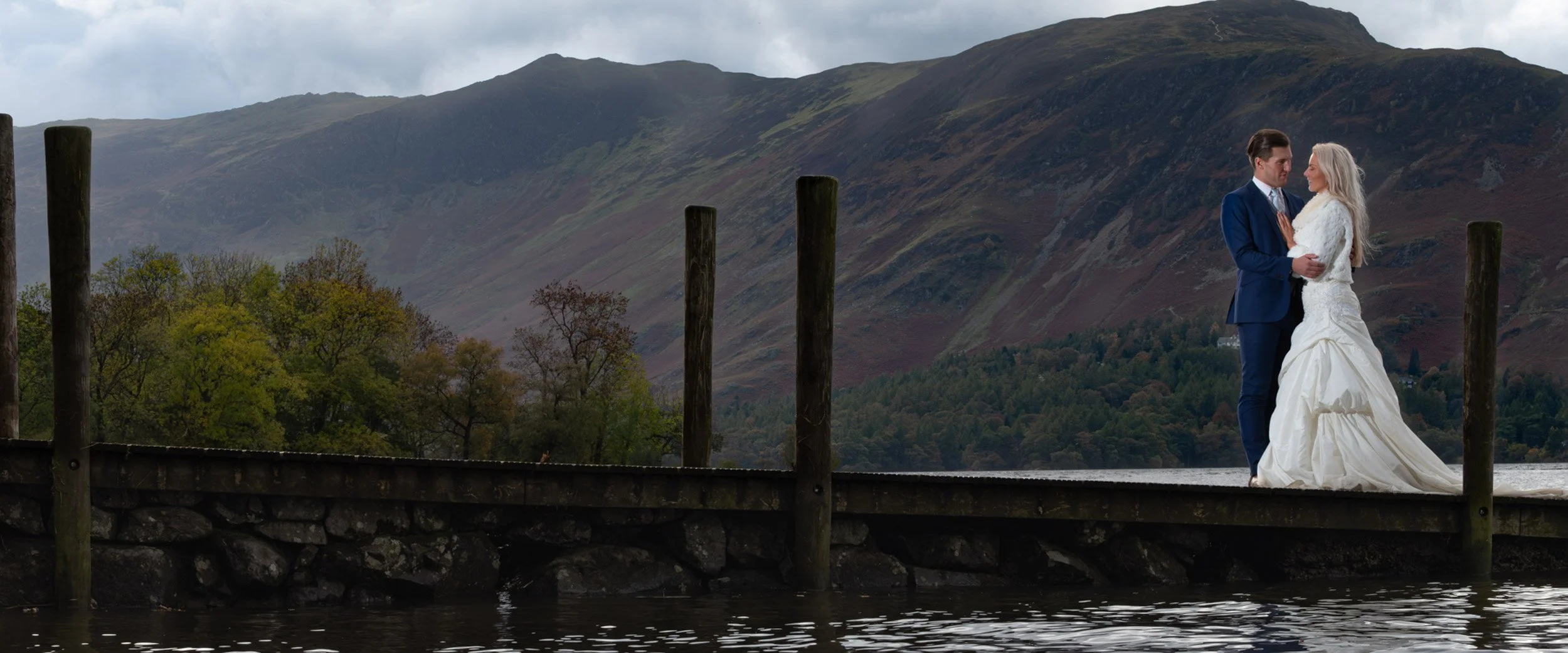A bride and groom standing close together on a wooden dock by a lake, with mountains and trees in the background during an overcast day.
