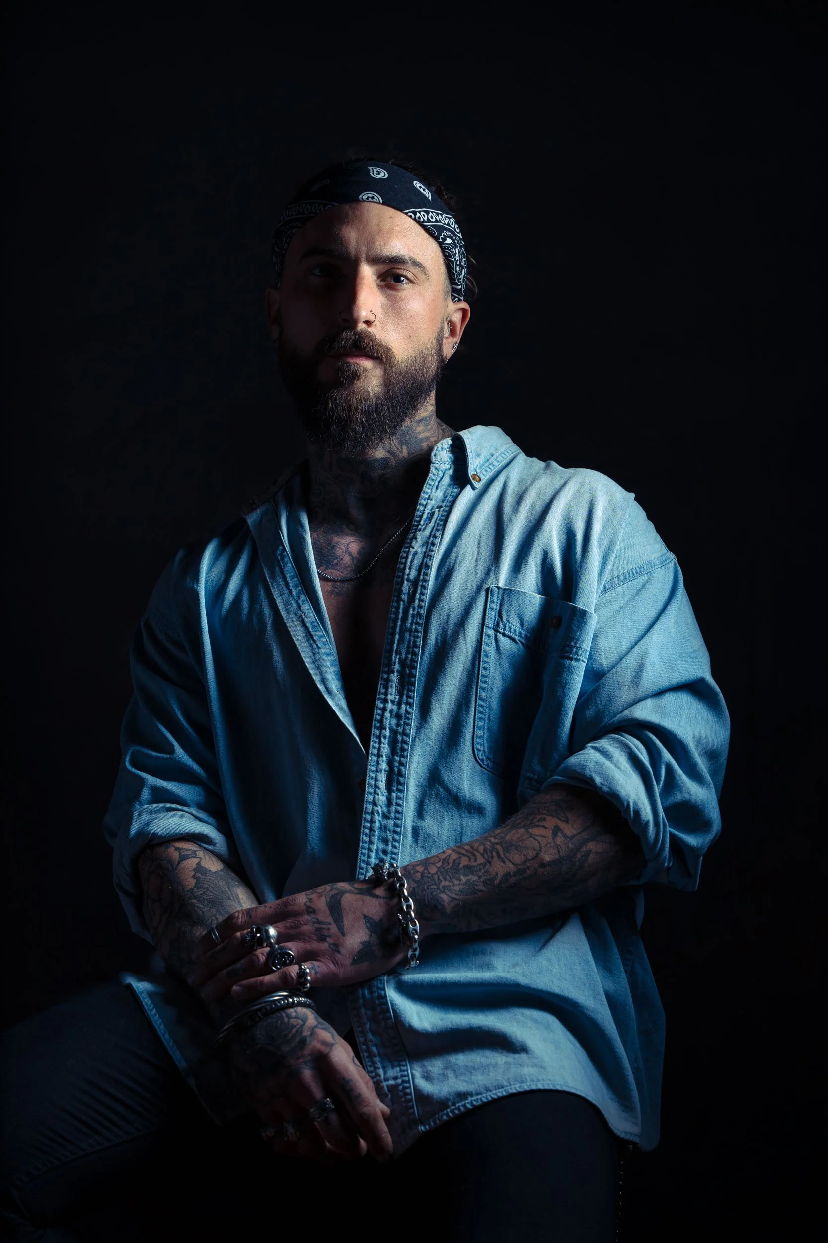 A man with tattoos, a beard, and a bandana, wearing a denim shirt, sitting against a dark background.