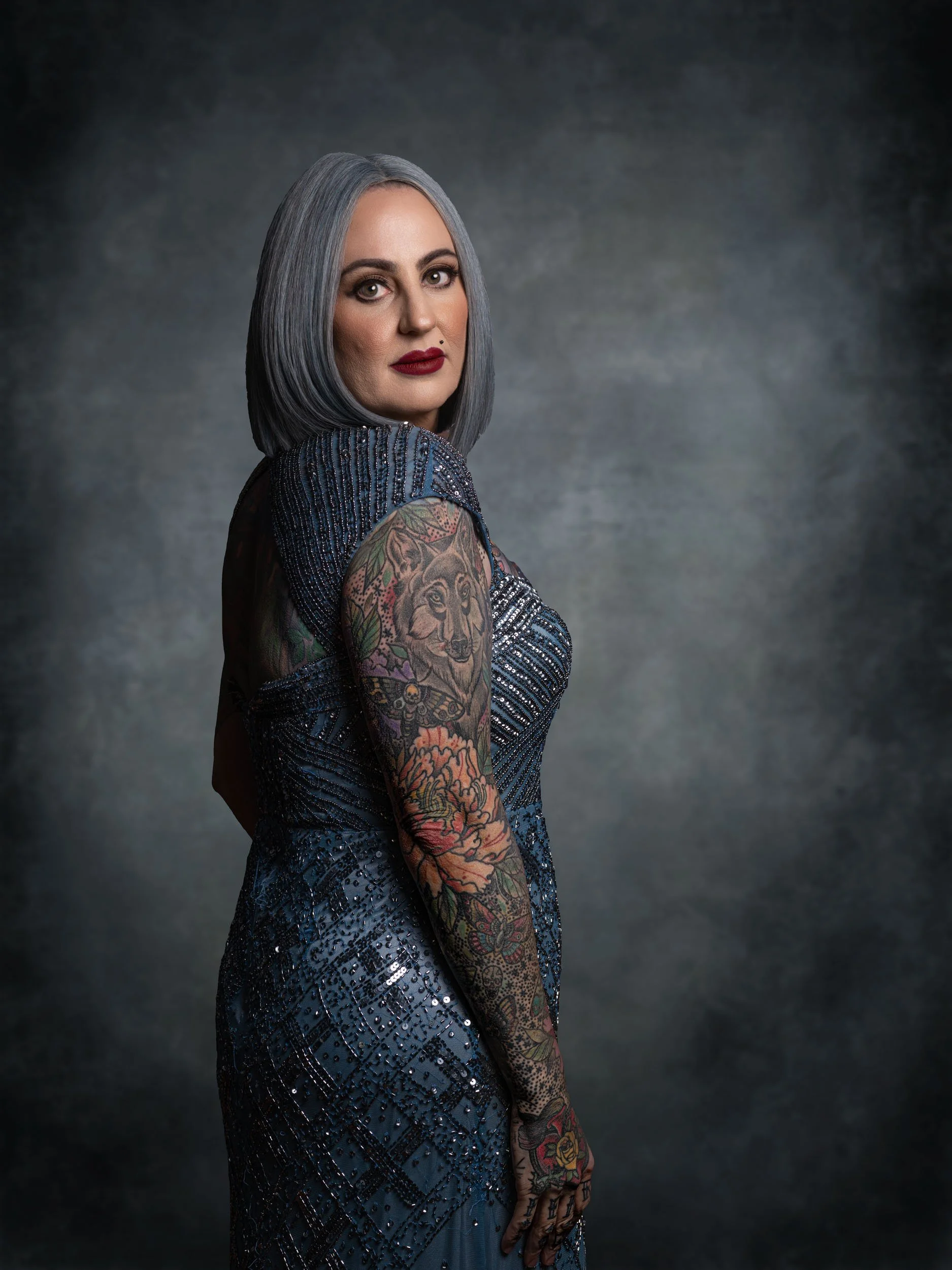 A woman with body tattoos, gray hair, red lipstick, wearing a dark, sparkly dress, standing against a dark background.
