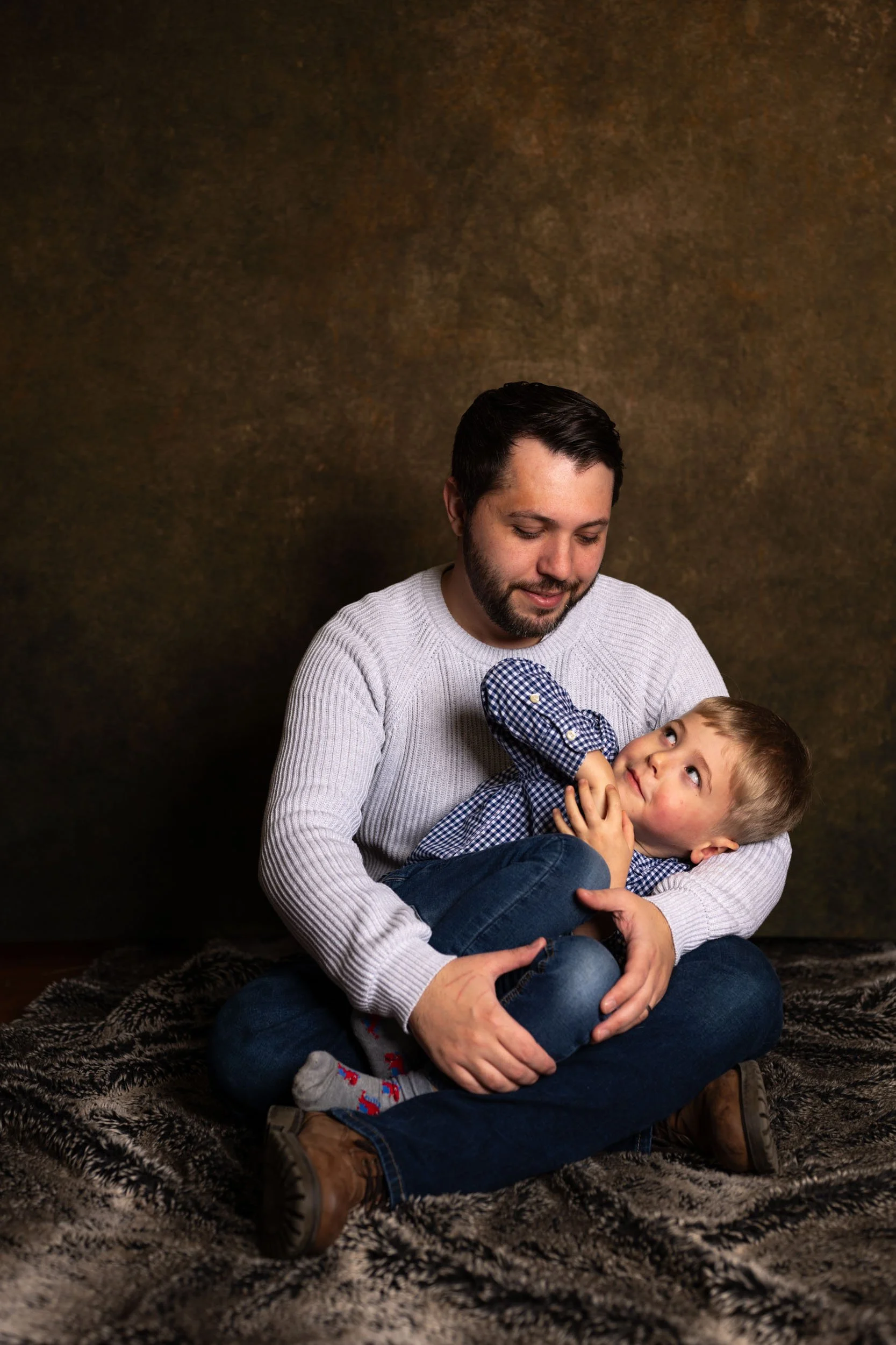 A man and a young boy sitting on a textured blanket against a brown backdrop. The man is smiling gently while the boy looks up at him with an affectionate expression, their faces close together. The boy is wearing a blue checkered long-sleeve shirt a