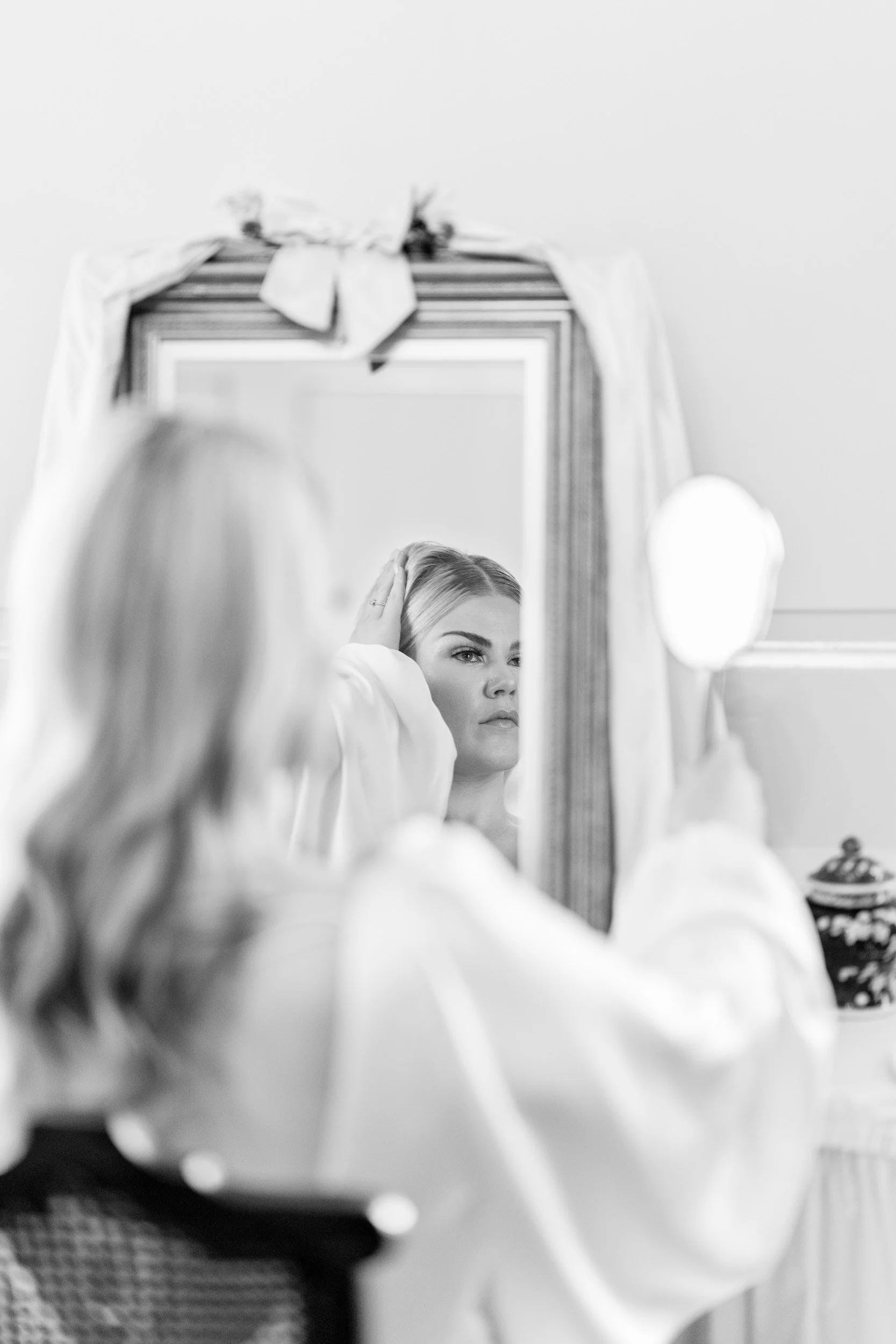 A woman looking into a mirror, adjusting her hair, reflected in a large framed mirror with a decorative bow on top, in a well-lit room.