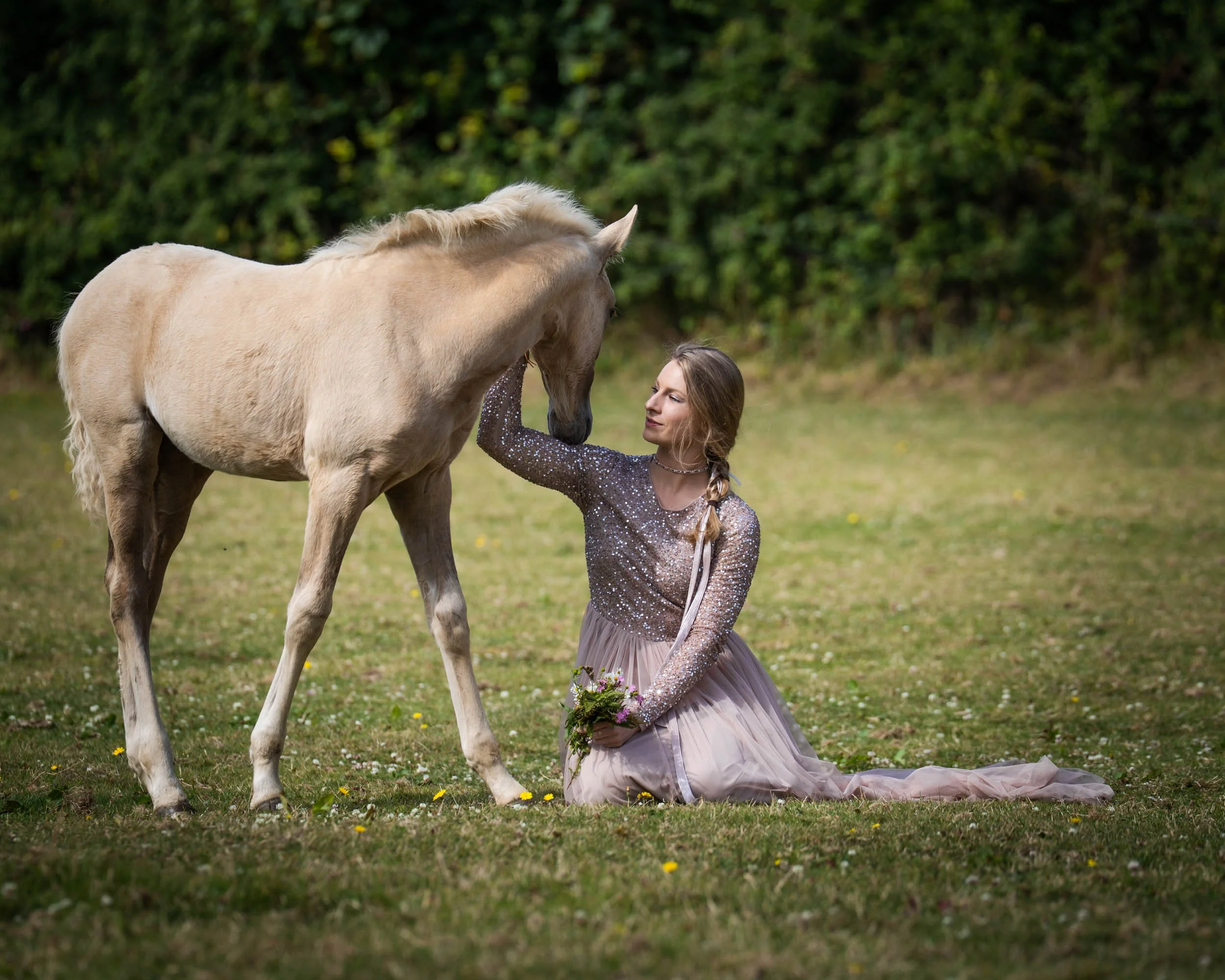 A woman in a sparkly long-sleeve dress kneels on grass, holding a bouquet, while gently touching a light-colored horse's face in a green outdoor setting.