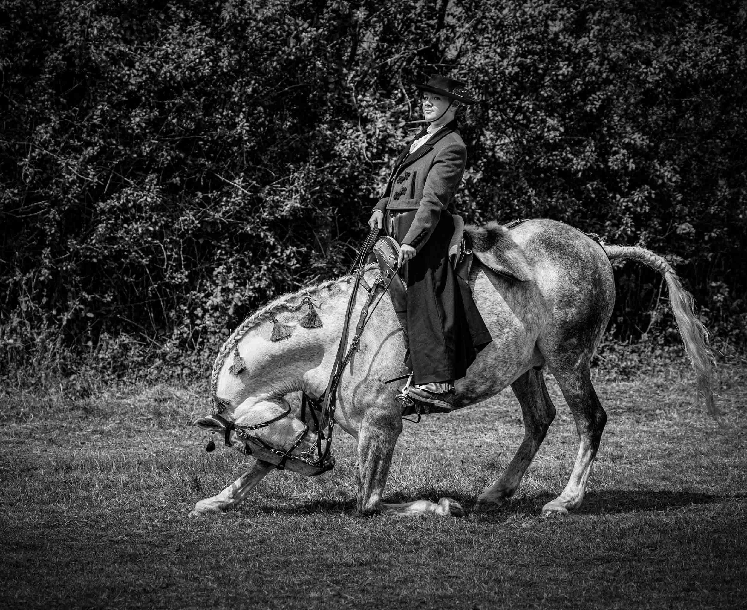 A woman in vintage clothing riding a horse, with her head turned slightly to the side, holding the reins. The horse is kneeling on the grass, and the background is filled with dense bushes or trees. The image is in black and white.