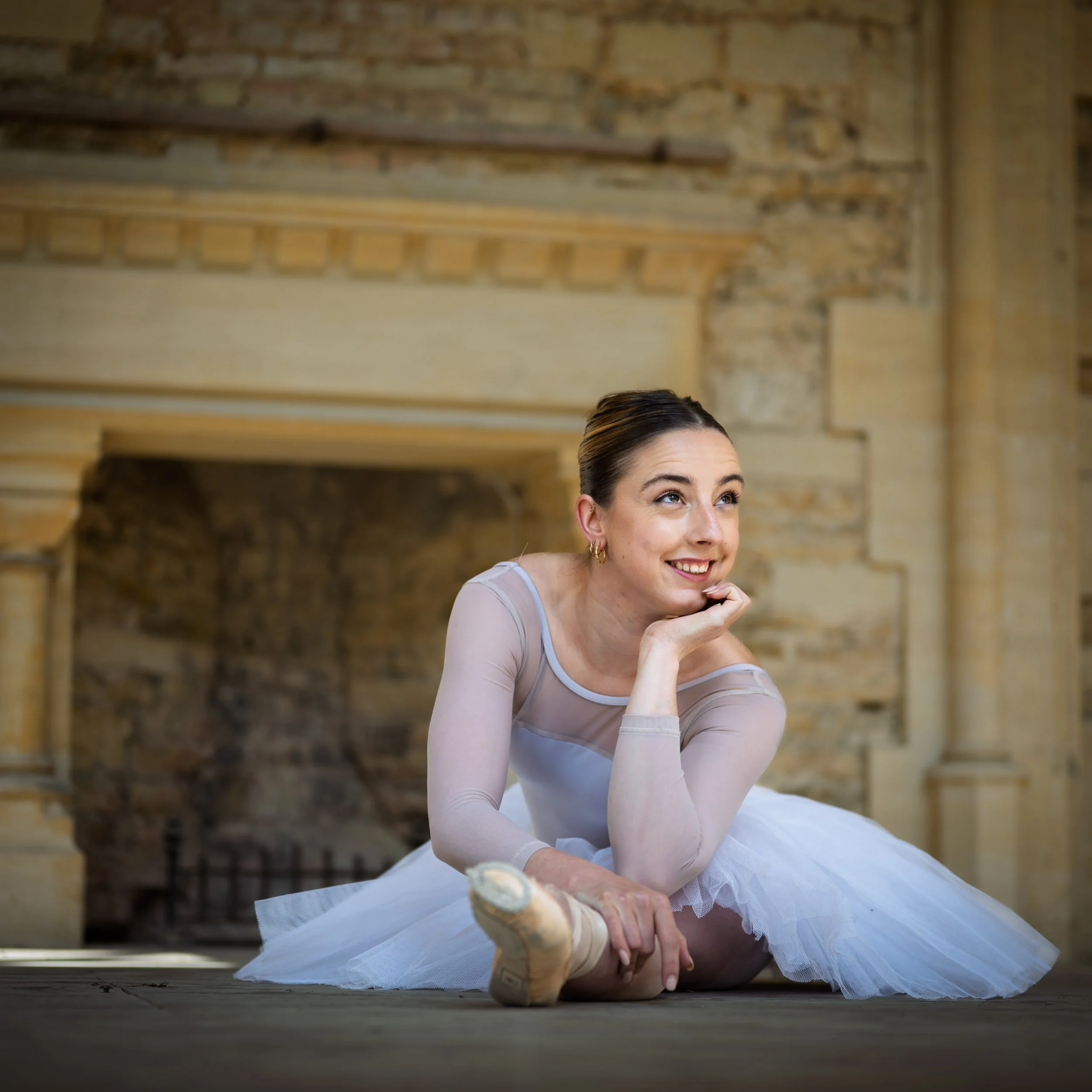 A ballerina in a tutu and ballet slippers sitting on the floor in a rustic, stone-walled room, smiling and looking up thoughtfully.