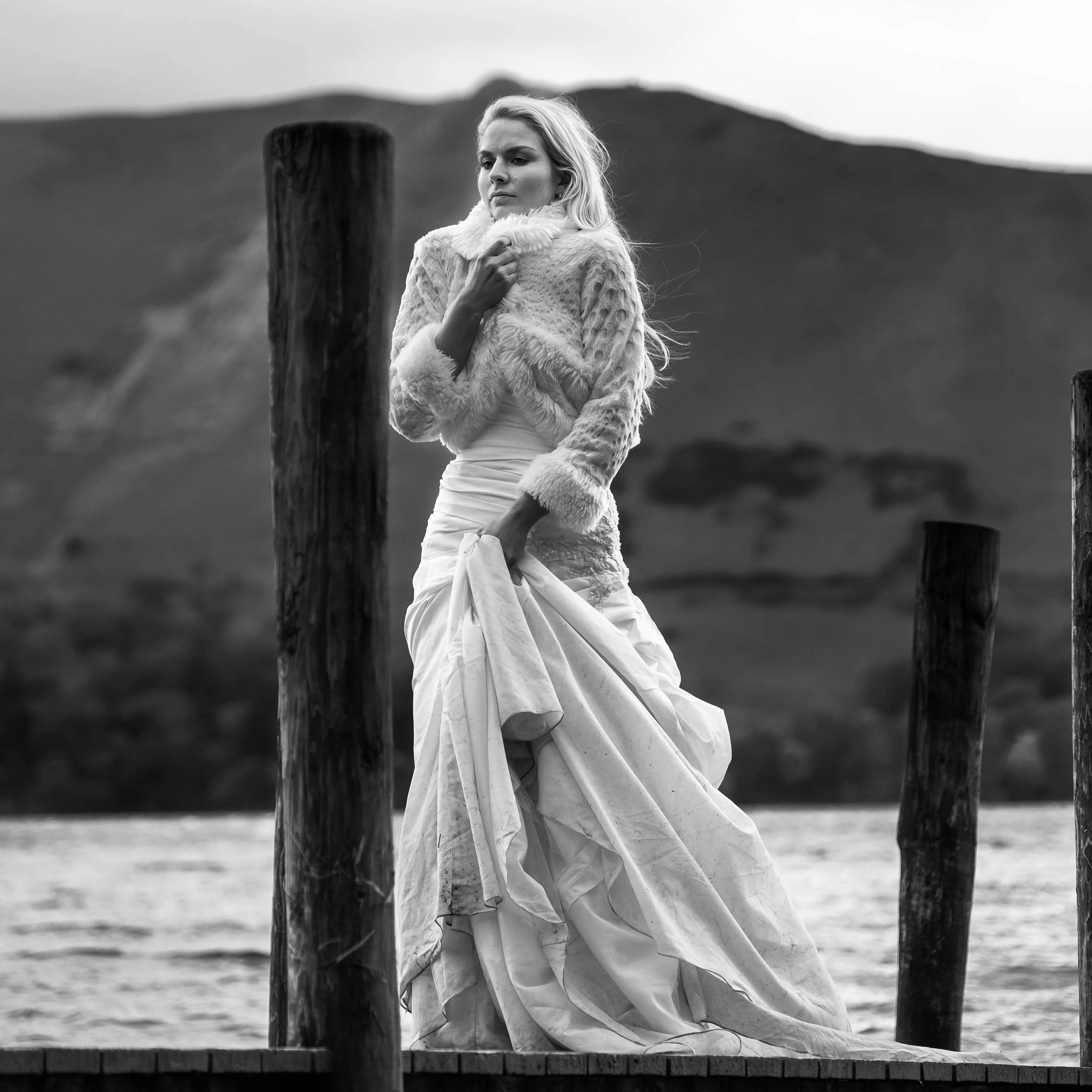 A black and white photograph of a woman in a wedding dress standing near water with mountains in the background, holding her dress and touching her collar with a pensive expression.