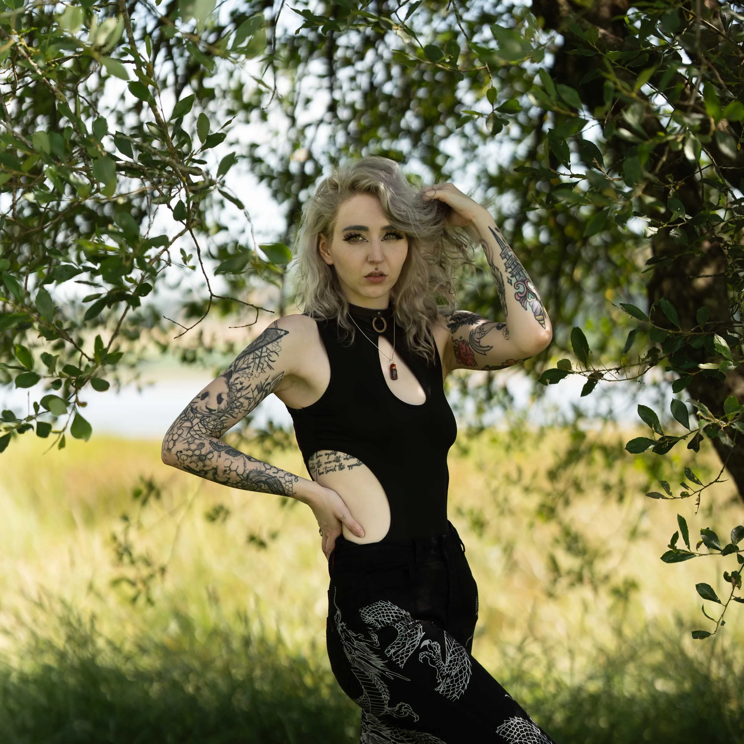A woman with tattoos on her arms and side, blonde wavy hair, and wearing a black open-side top and black pants with a dragon pattern, standing outdoors among green trees and grass.