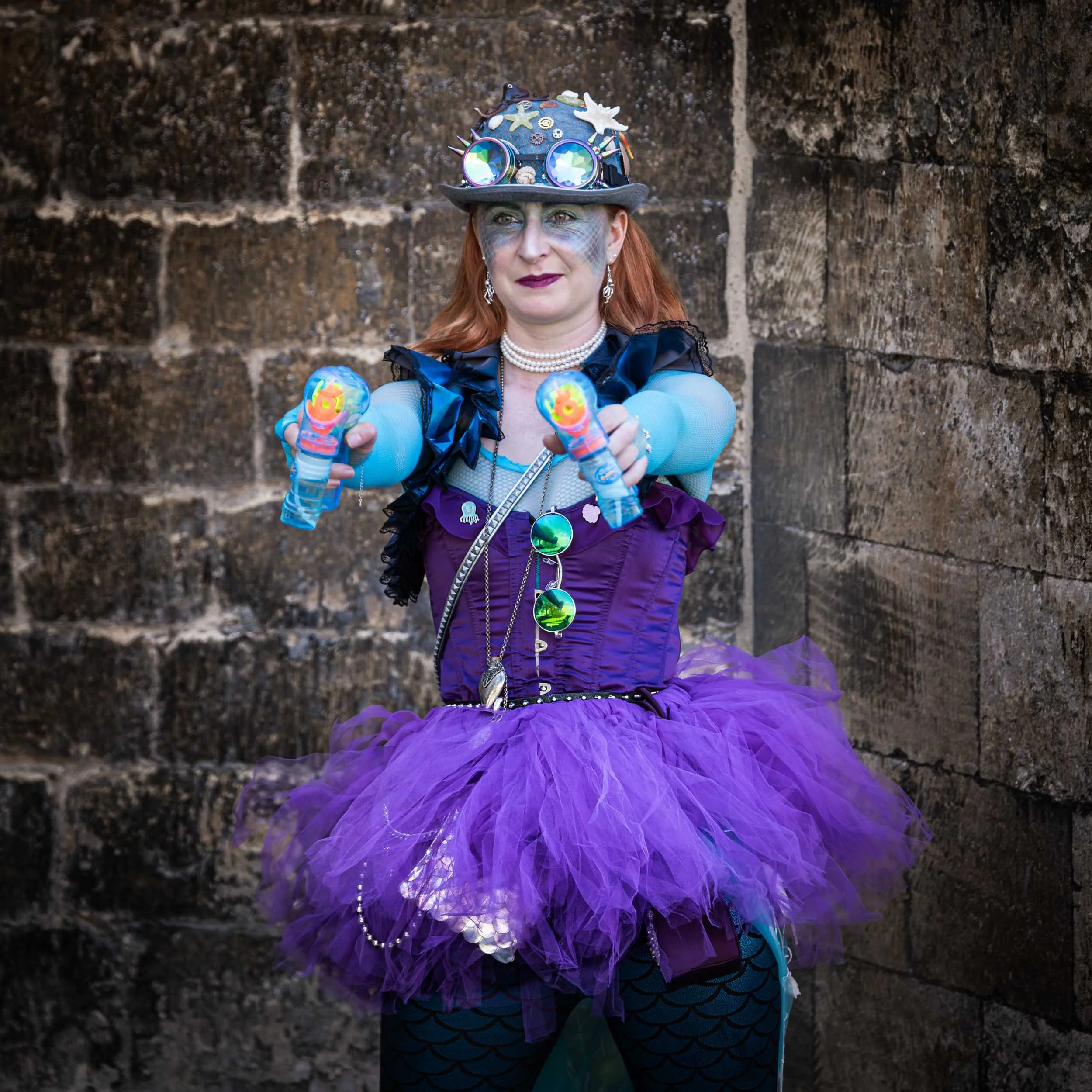 A woman with red hair dressed as a whimsical character in a purple tutu and mermaid scale leggings, wearing a decorated hat, pointing water guns at the camera, standing against a stone wall.