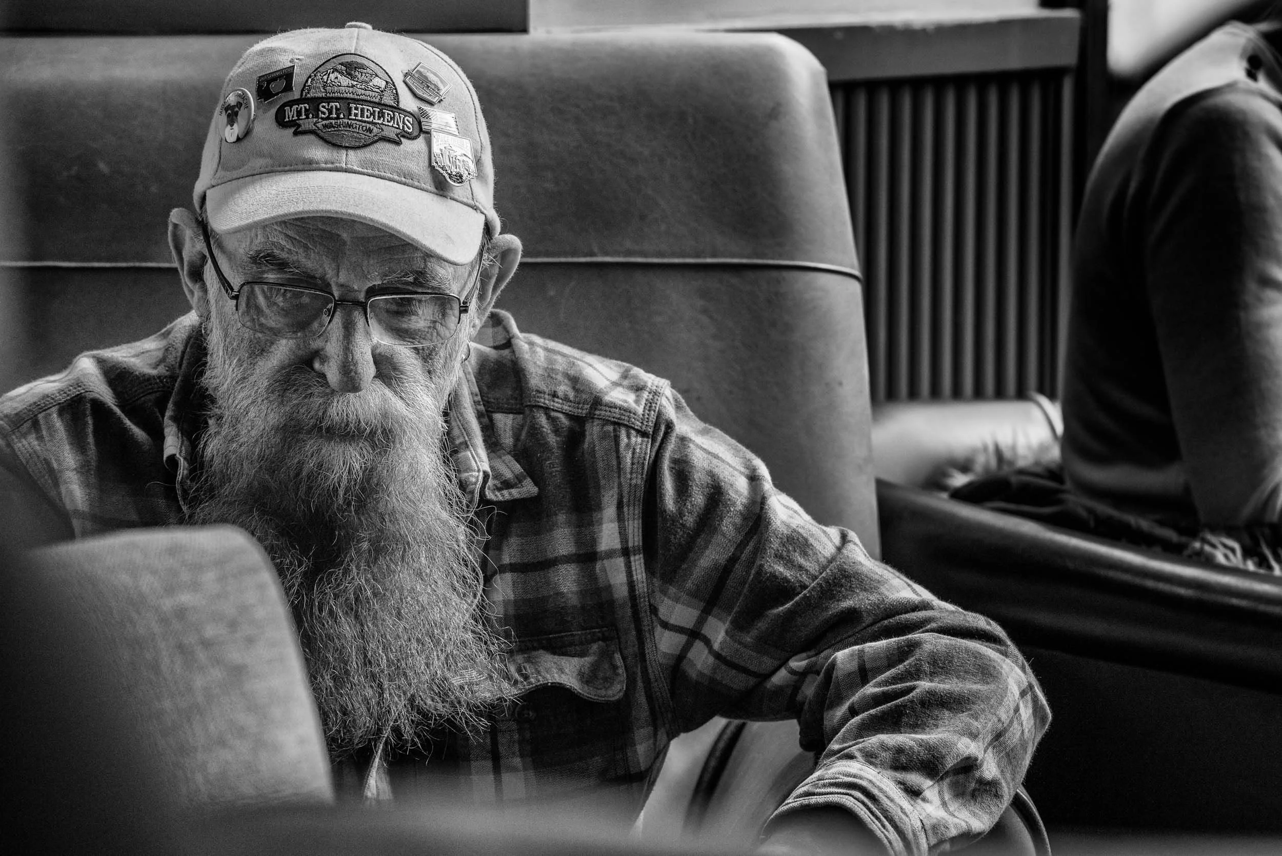 An elderly man with a long beard, glasses, wearing a plaid shirt and a cap with patches, sitting and looking down.