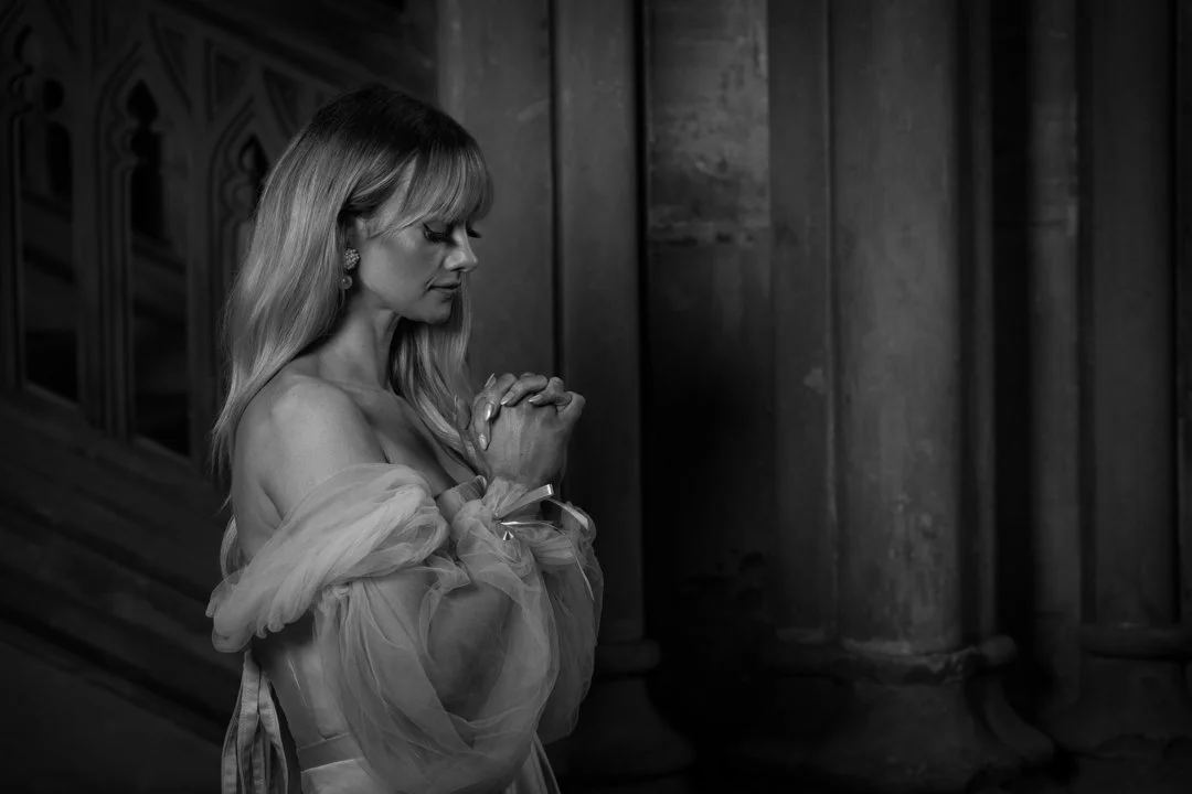 A woman with long blonde hair and earrings, dressed in a sheer, off-shoulder gown, standing with her hands clasped in prayer inside a church or cathedral with tall stone columns.