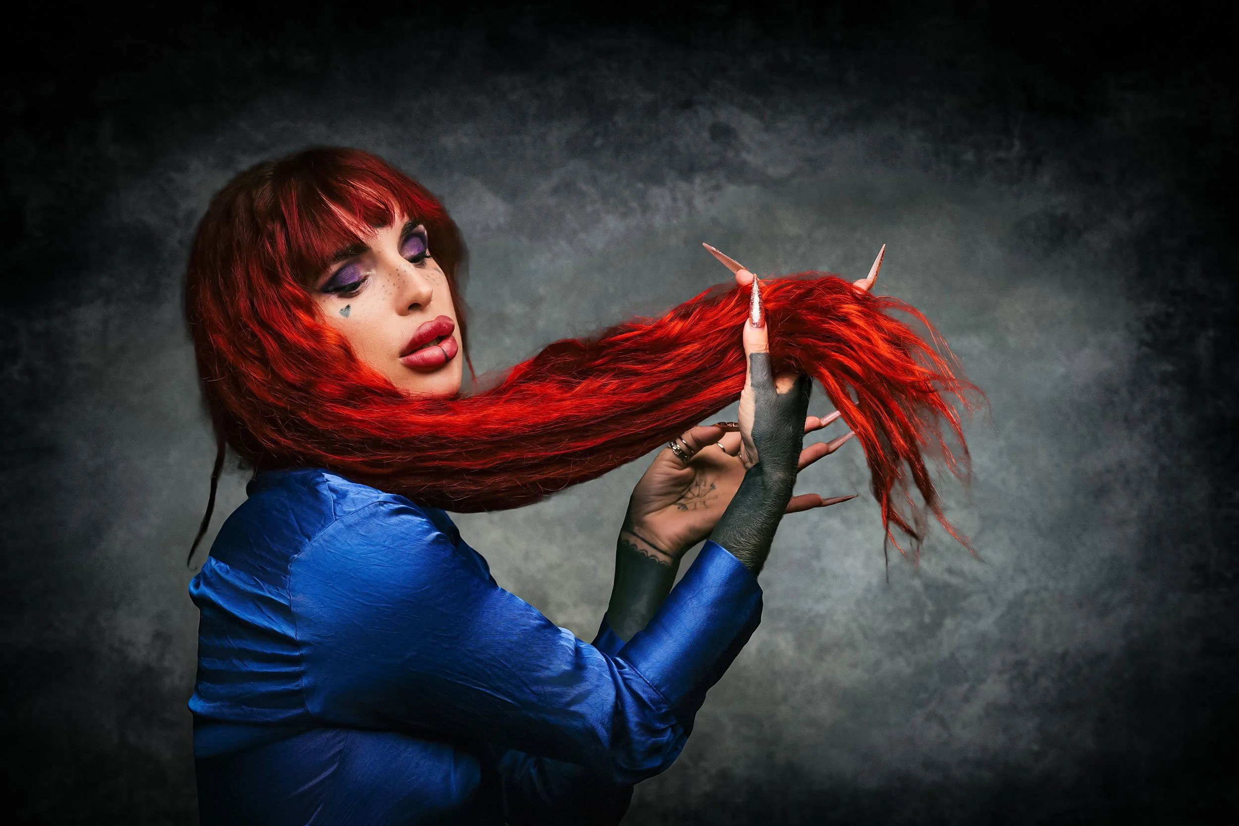 A woman with long red hair and platinum blonde roots, wearing a blue satin jacket, holding her hair with her left hand, which has painted nails and rings, against a dark textured background.