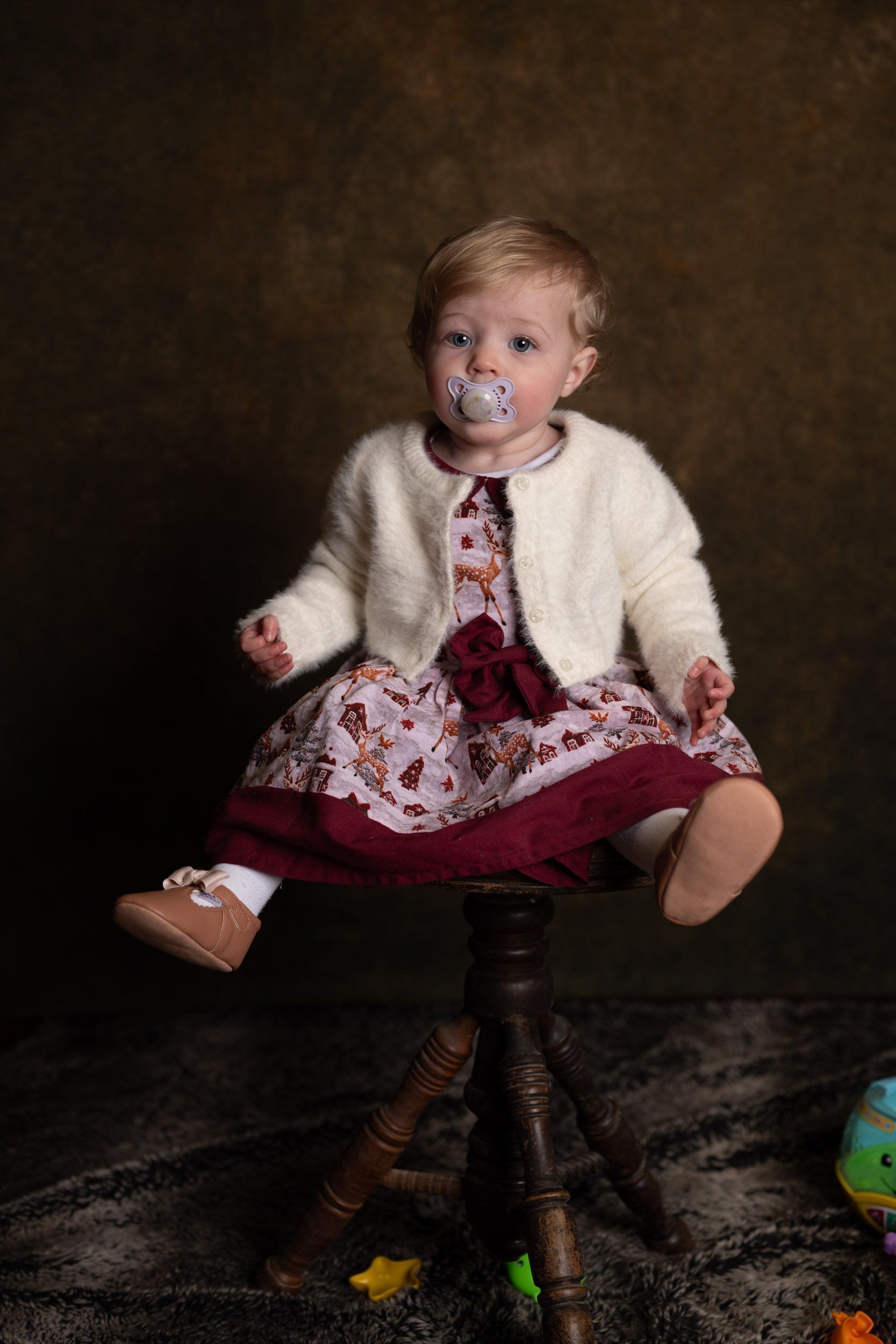 A young child with light brown hair and blue eyes sitting on a wooden stool, wearing a festive dress with reindeer and snowflakes, a cream-colored cardigan, and light brown shoes with white socks. The child has a pacifier in their mouth and is lookin
