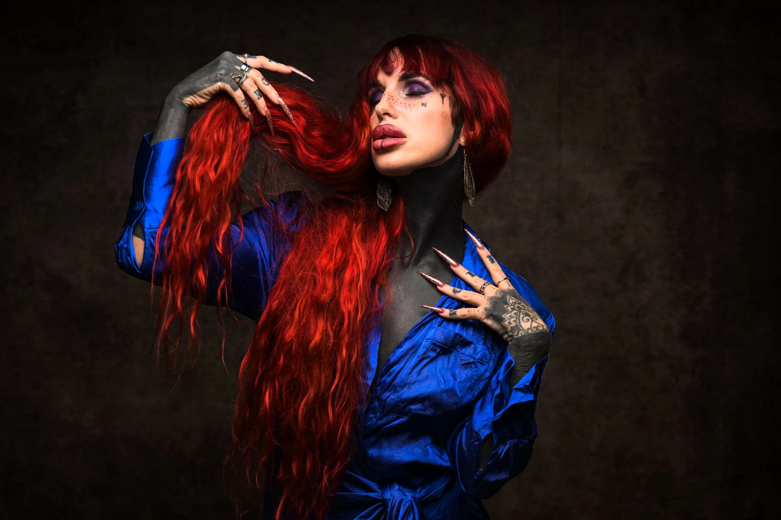 A woman with long, wavy red hair, makeup, and tattoos on her hand and face, wearing a blue jacket with a black turtleneck, and posing with her eyes closed against a dark background.