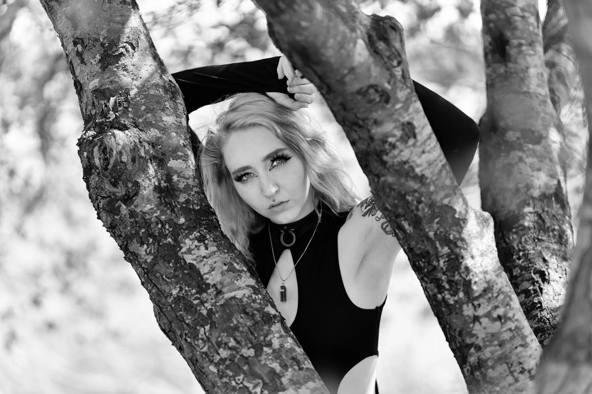 Black and white photo of a young woman with wavy hair and tattoos, posing outdoors between tree branches, looking at the camera with a serious expression.