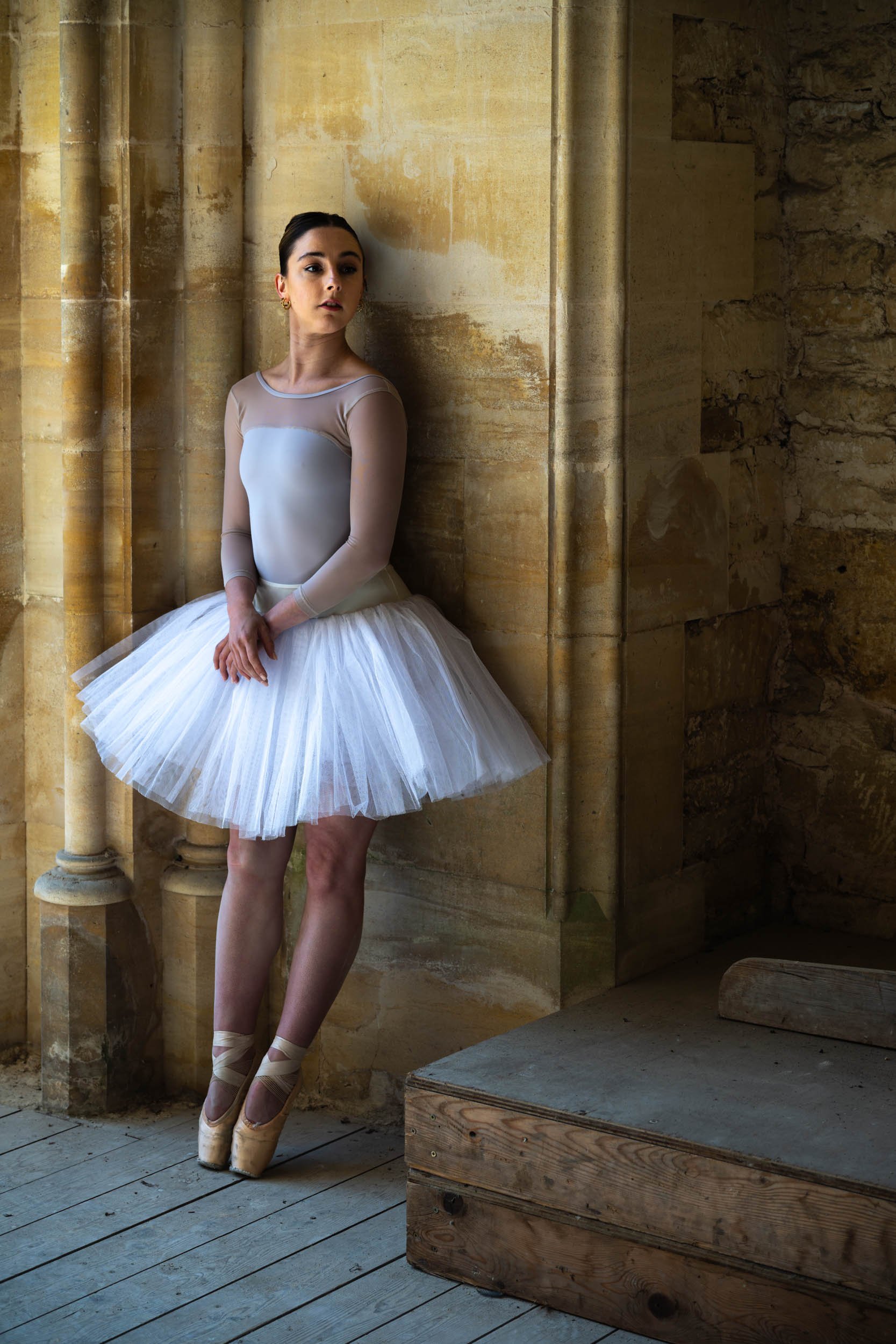 A ballerina in a white tutu and ballet shoes standing against a stone wall in a ballet studio.