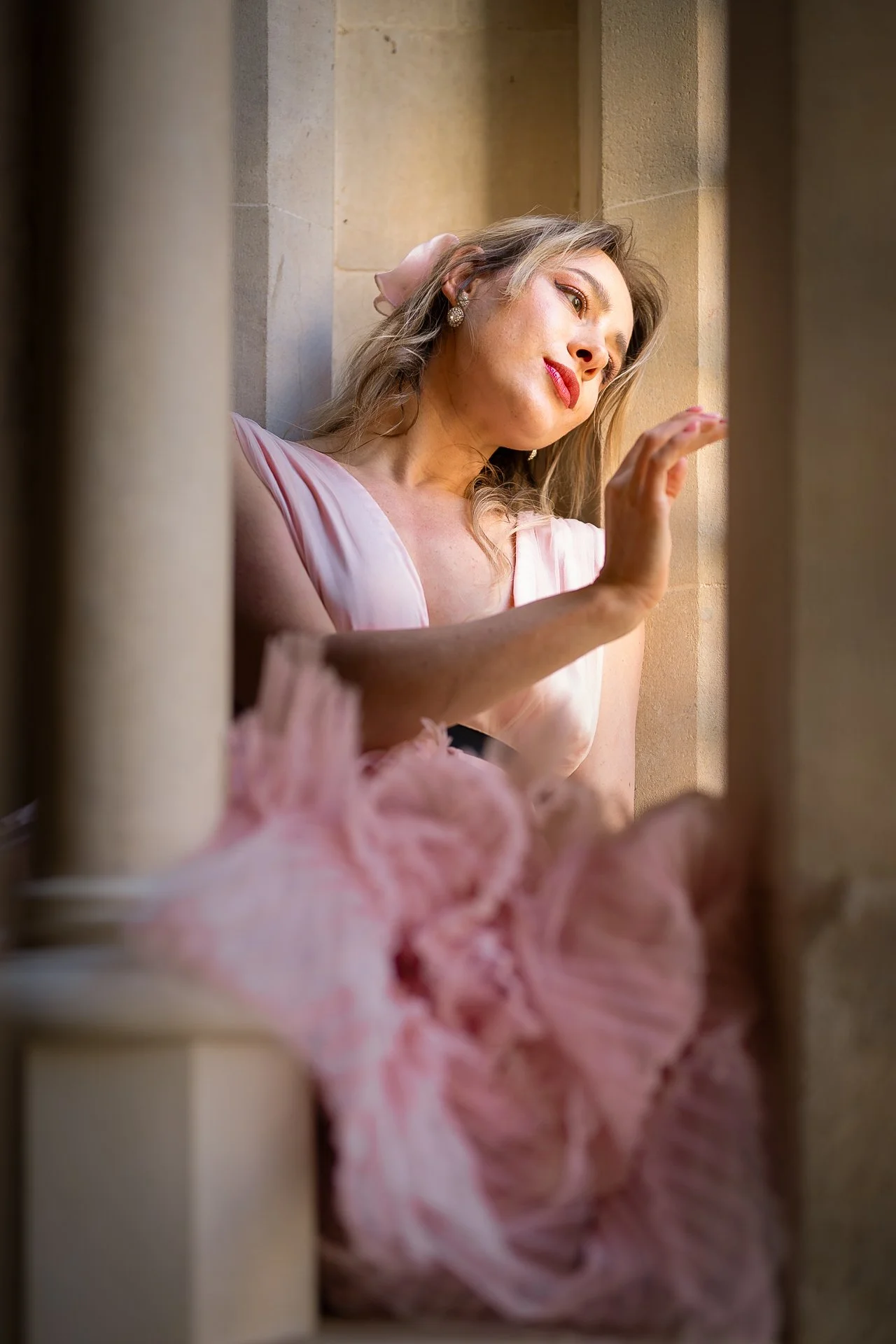 A woman in a pink dress reclining against a beige stone wall, looking peacefully to the side with her hand raised near her face.