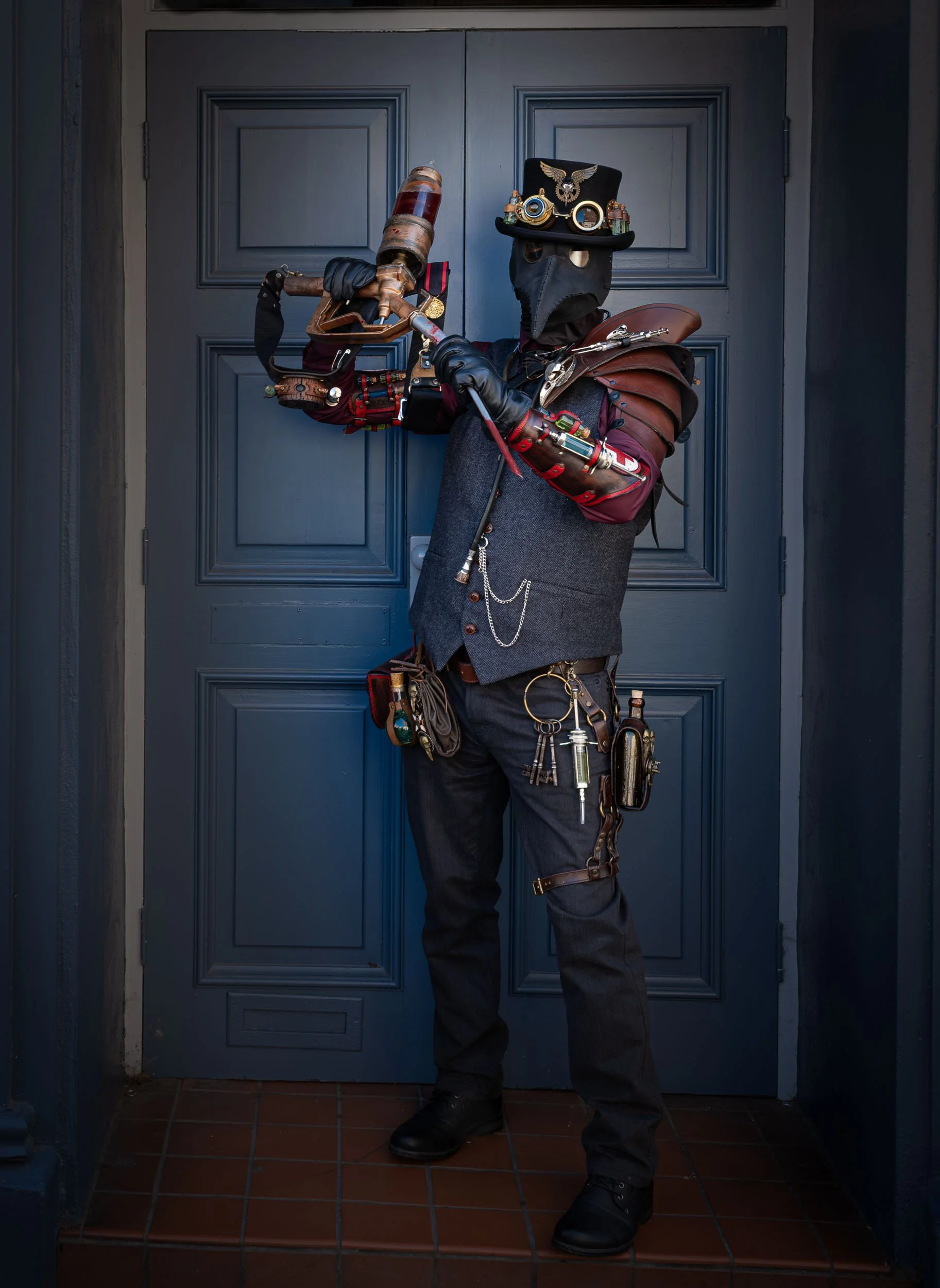 Person dressed in steampunk costume with goggles, gears, and mechanical accessories, holding a weapon, standing in front of a blue door.