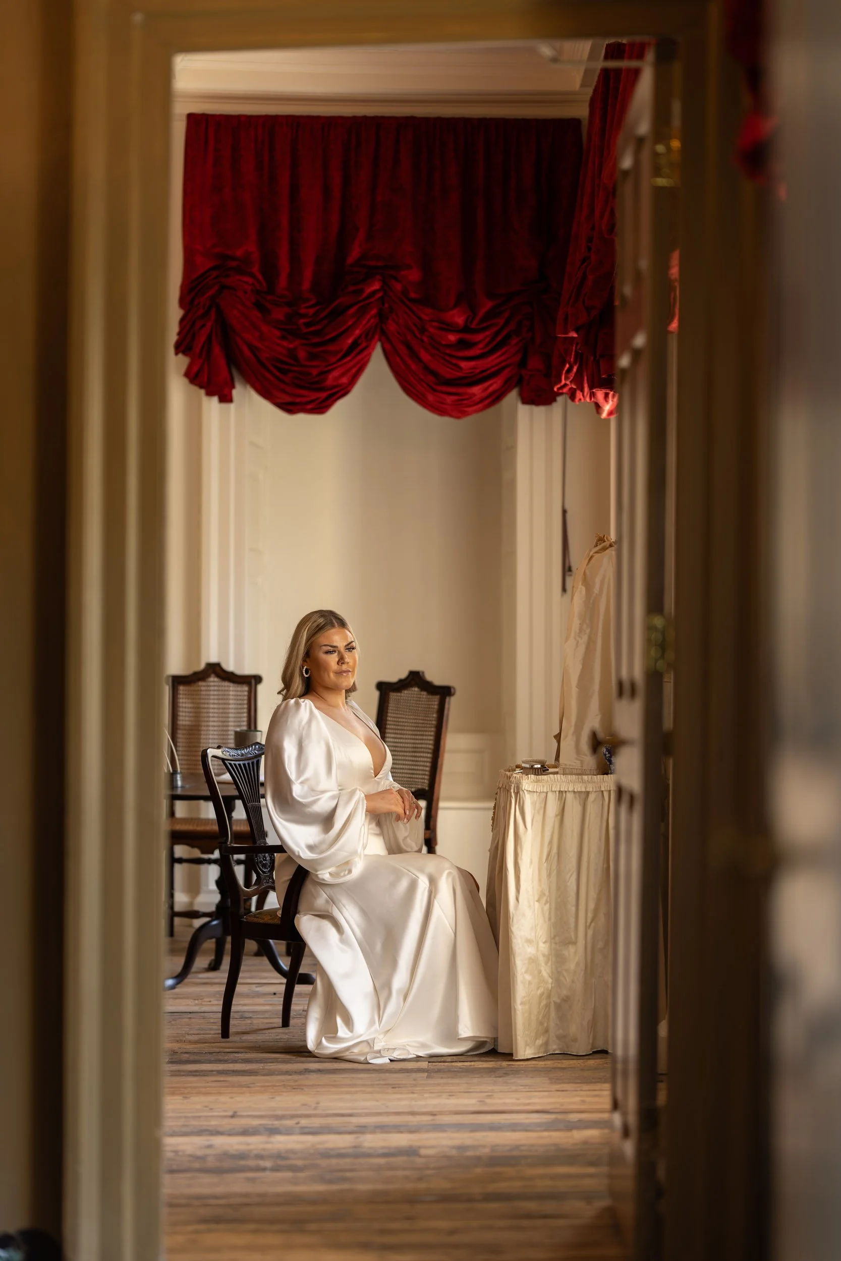 A woman in a white satin dress sitting at a vanity with a mirror, seen through a doorway with red drapes overhead.