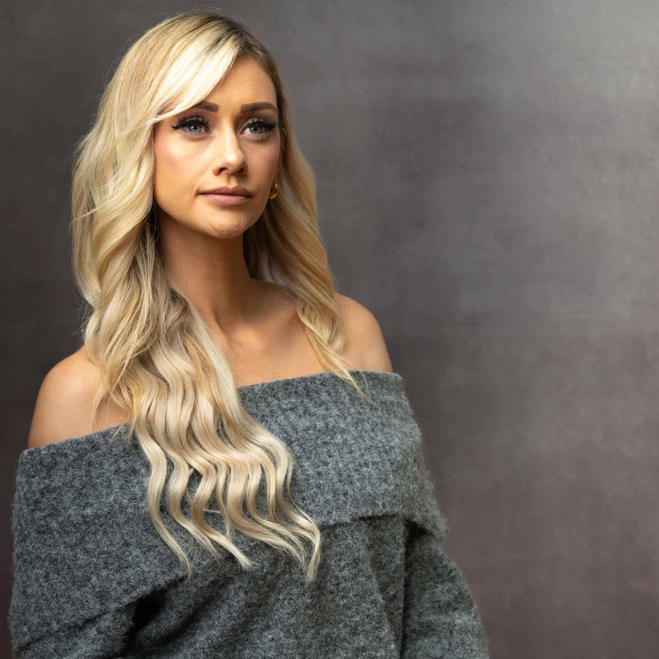 A woman with long blonde wavy hair, wearing an off-the-shoulder gray sweater, standing against a plain gray background.