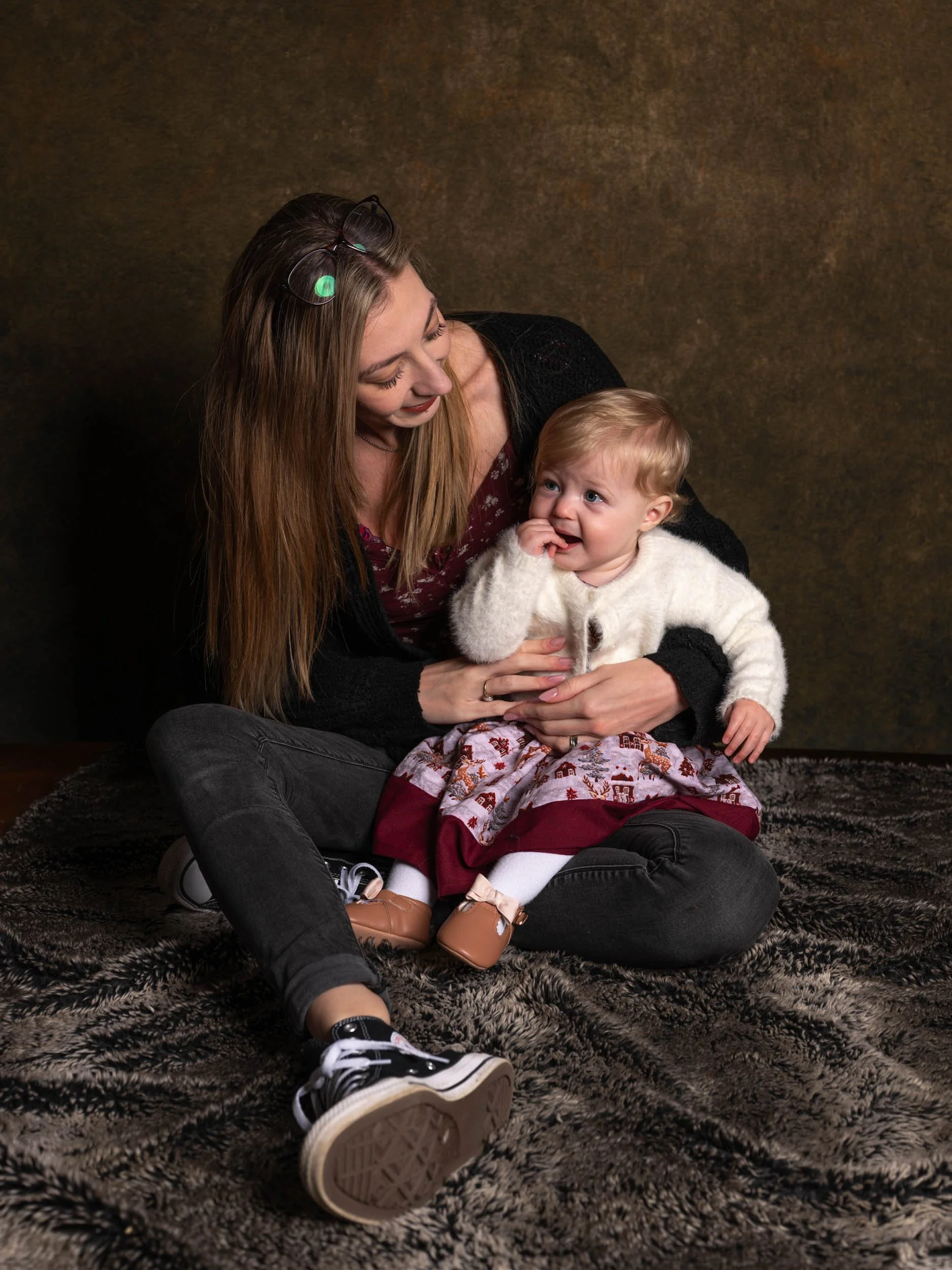 A woman with long blonde hair and a young girl sitting on a textured rug against a warm, dark background. The woman is holding the girl, who is wearing a white sweater, a festive skirt, and tan shoes. The girl has blonde hair and blue eyes, and is sm